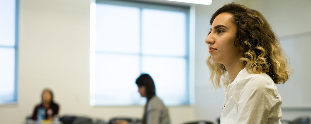 Girl in classroom pictured in profile. She has a long-sleeved white shirt, olive skin, and shoulder length curly light brown hair.