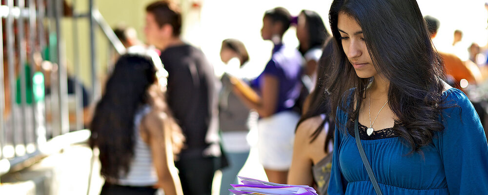 Female student with long straight black hair looks at some papers in her hand. She wears a bright blue long-sleeved shirt.