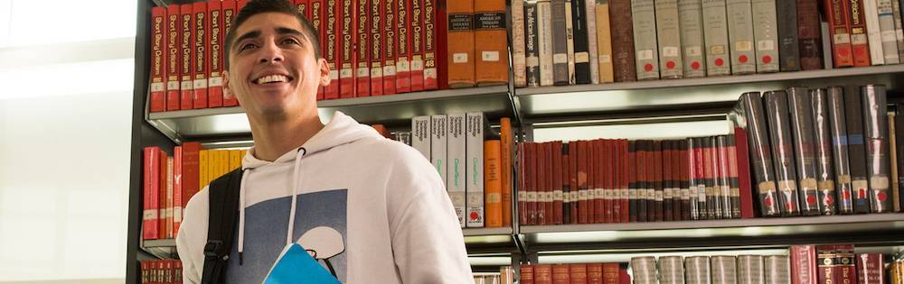 Lantino college student with short black hair, white tshirt, and books under arm smiles. He stands in front of library shelves.