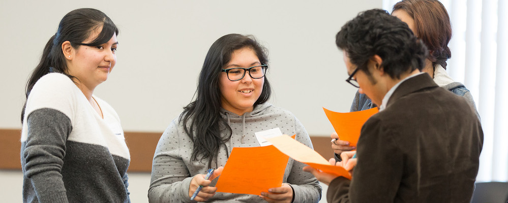 Three students of LatinX descent hold bright folders in a classroom. 