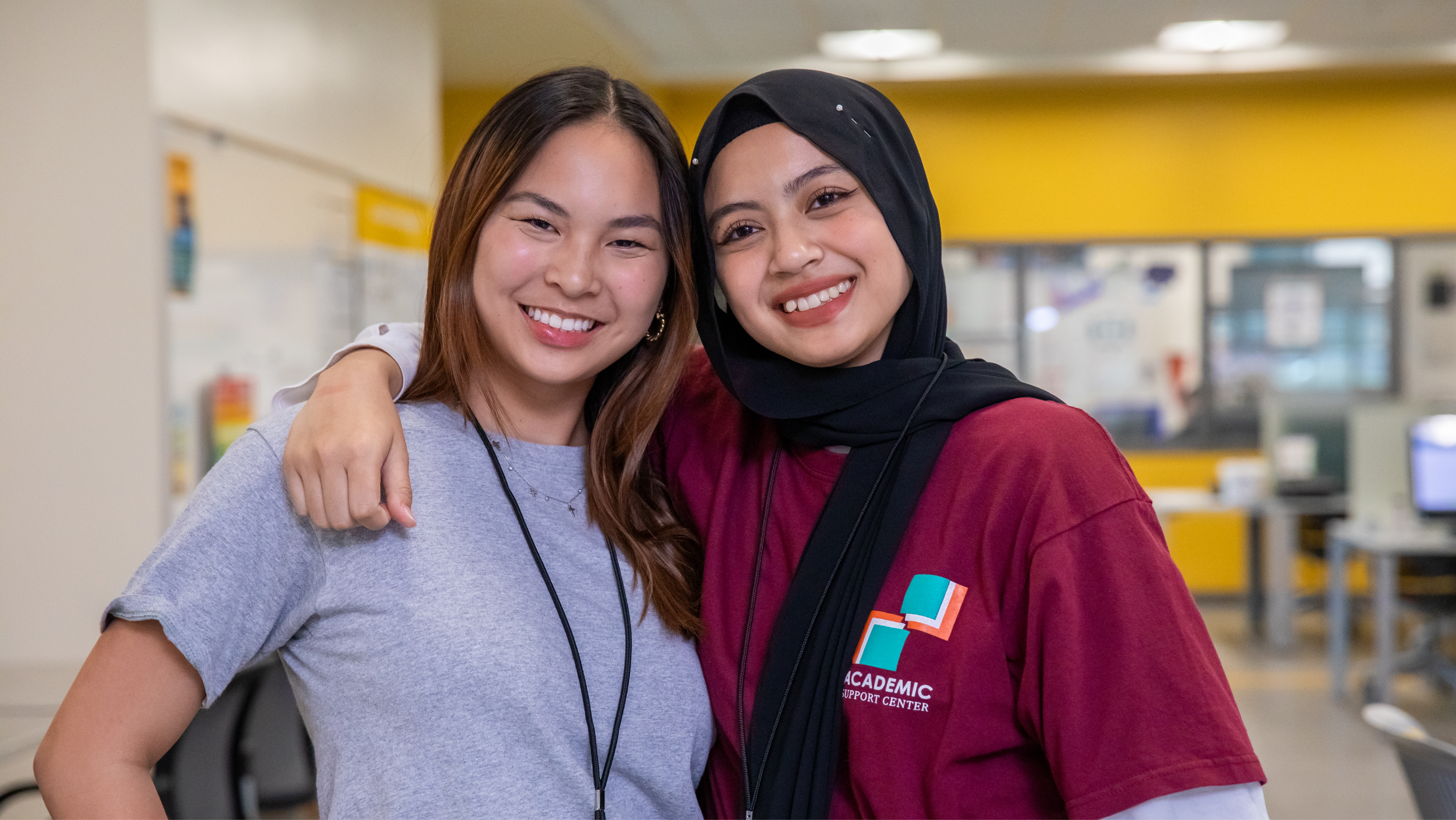 Two female students smiling closely together with arms around each other's shoulders; one wears a grey t-shirt and the other wears a black hijab and a maroon Academic Support Center t-shirt.