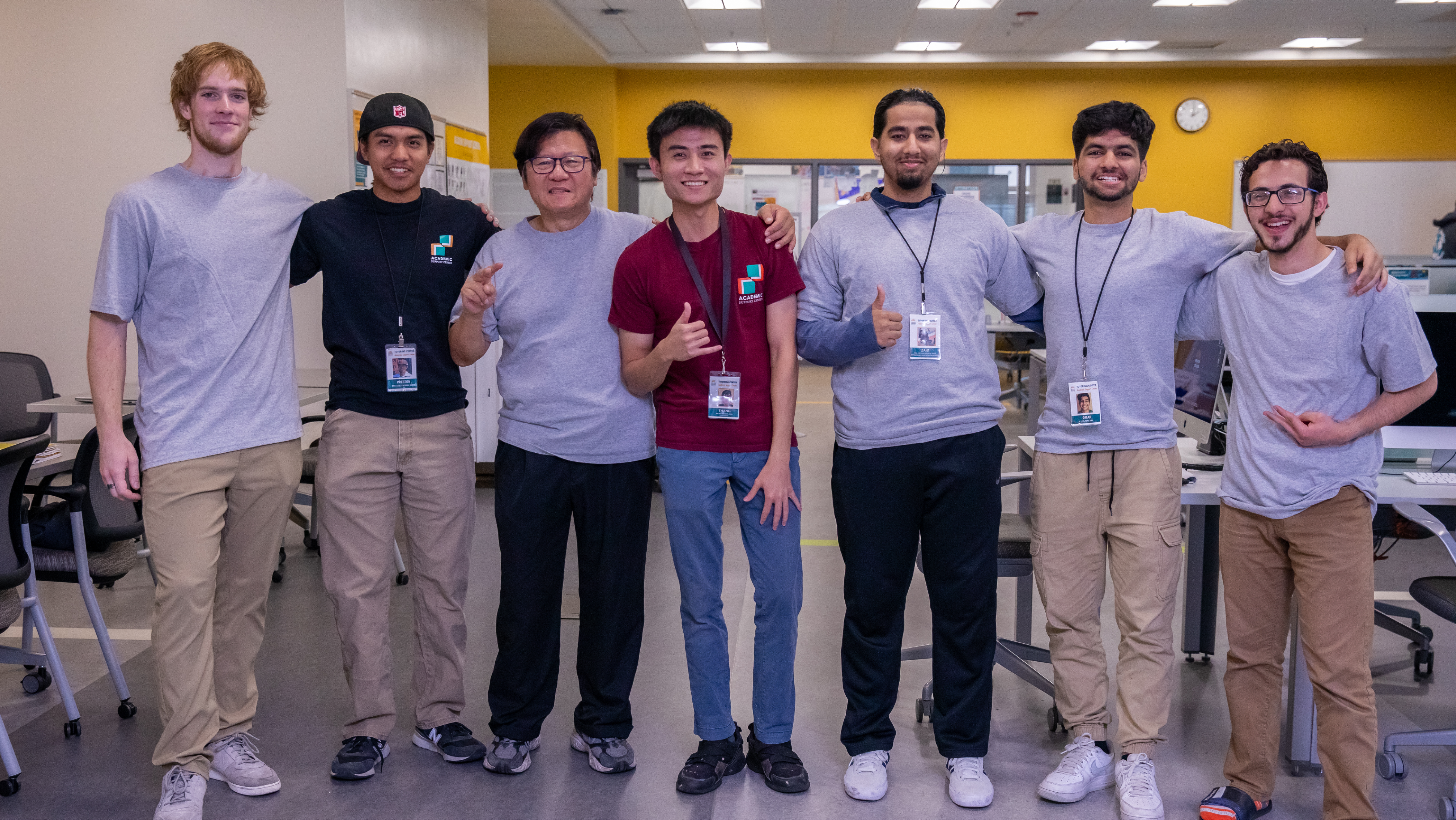 A group of seven young men standing arm-in-arm in an academic center, smiling at the camera with several wearing staff lanyards and Academic Support Center t-shirts.