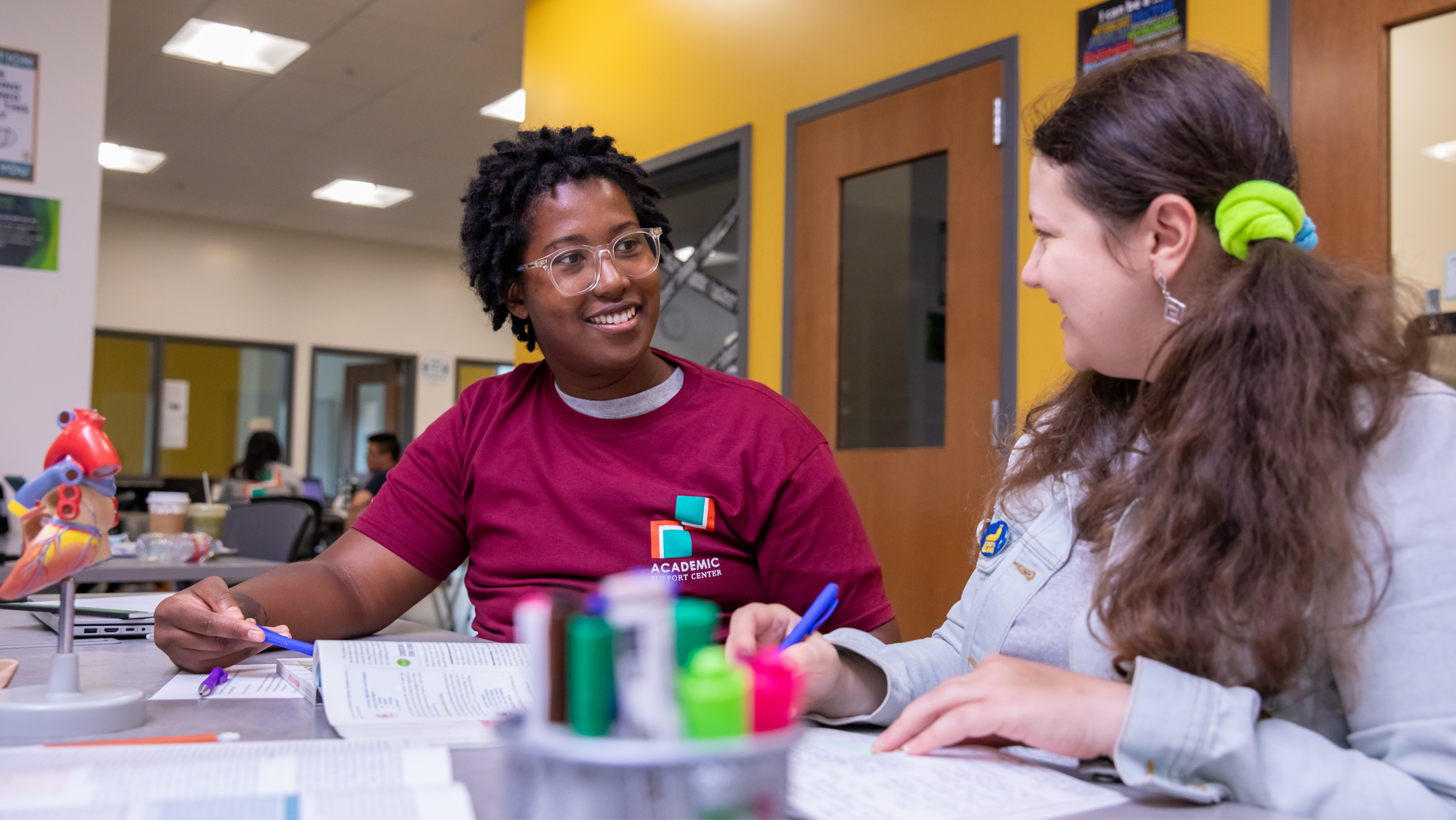 Two smiling students sitting at a table in a study center, pointing at an anatomical heart model while working together with open textbooks and a notebook.