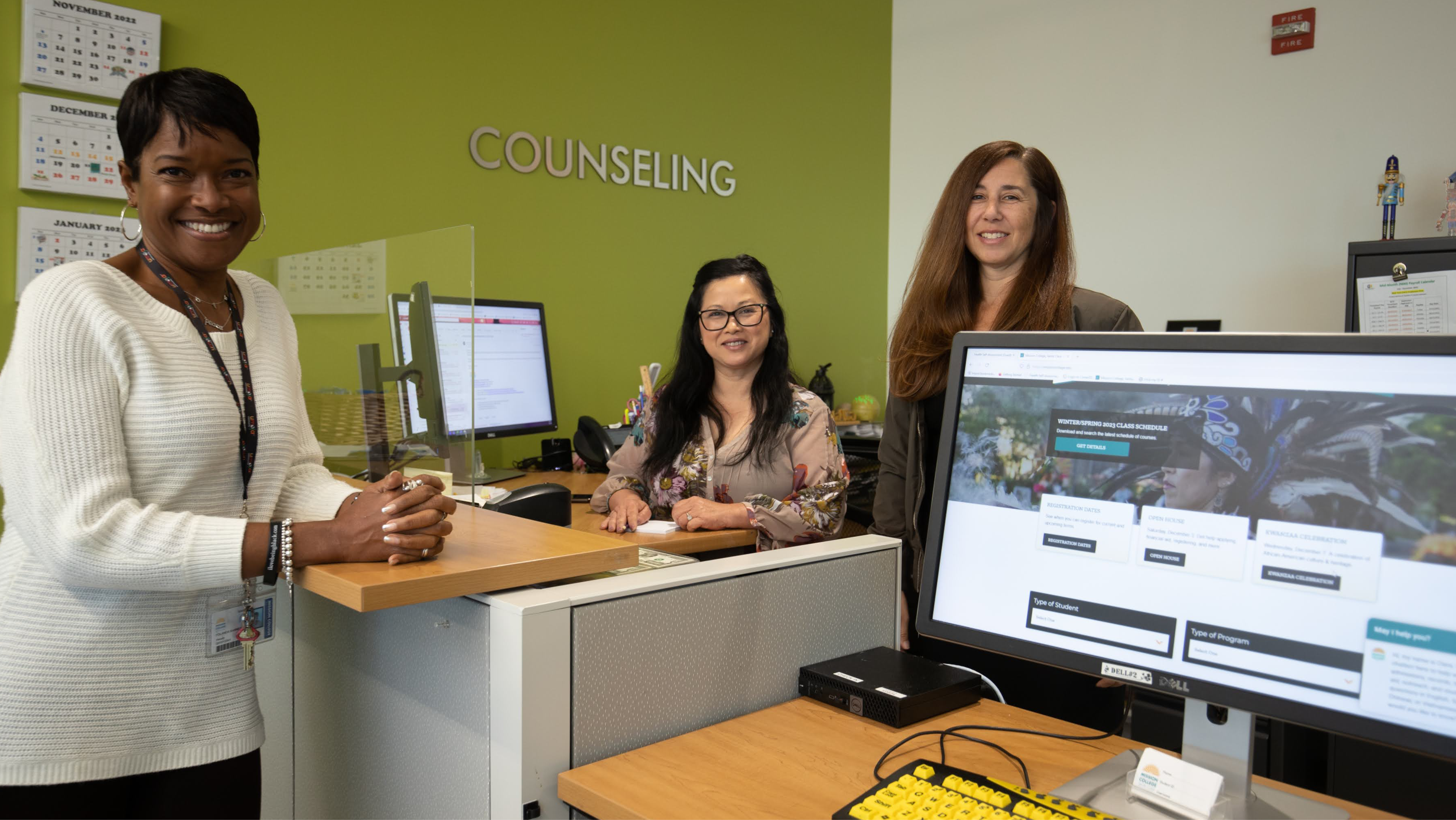 Three Mission College counseling staff members smiling behind the reception counter, with a computer screen displaying the college website in the foreground.