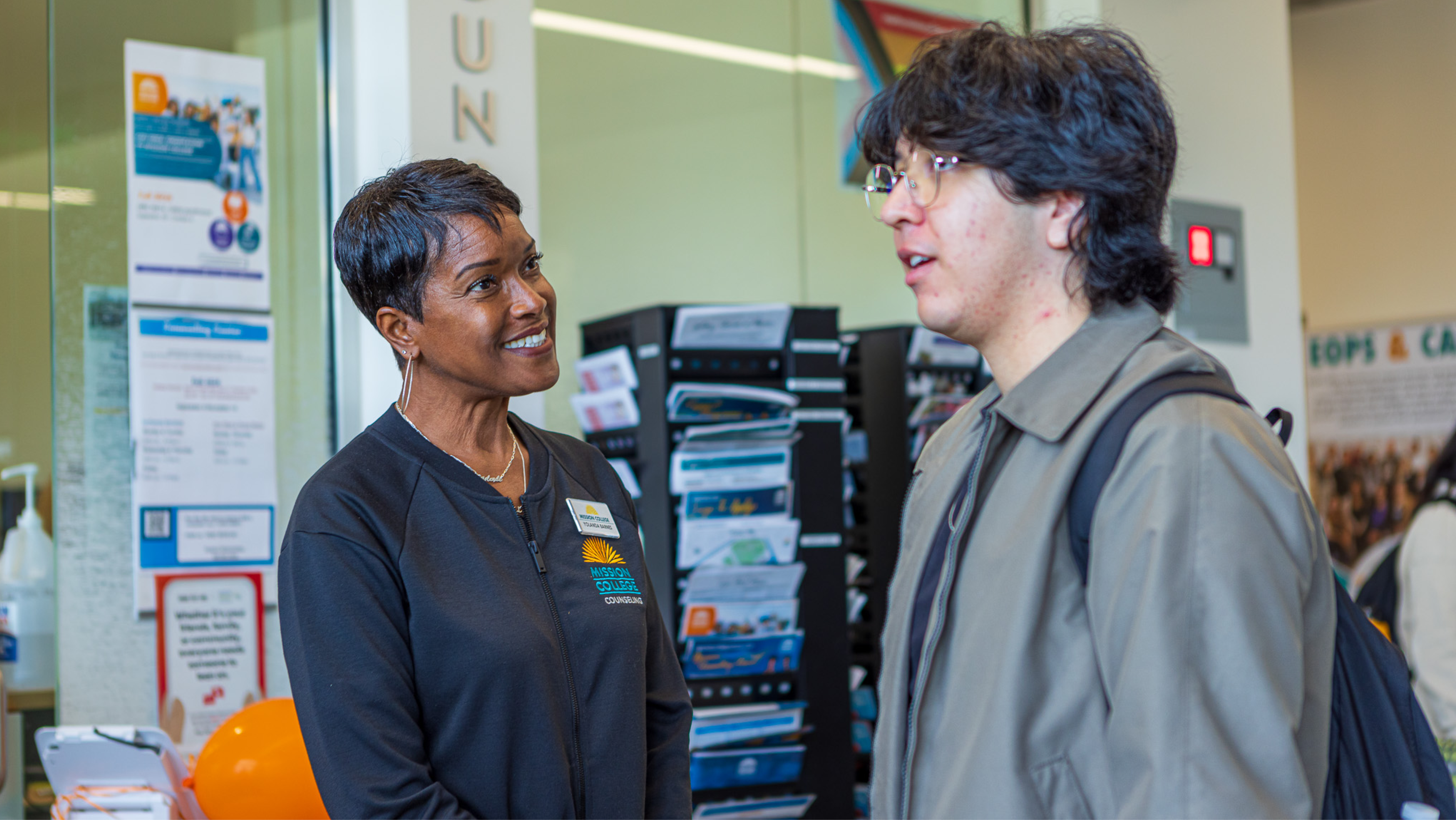 A female counselor in a black Mission College Counseling jacket smiling while listening to a student in the lobby.
