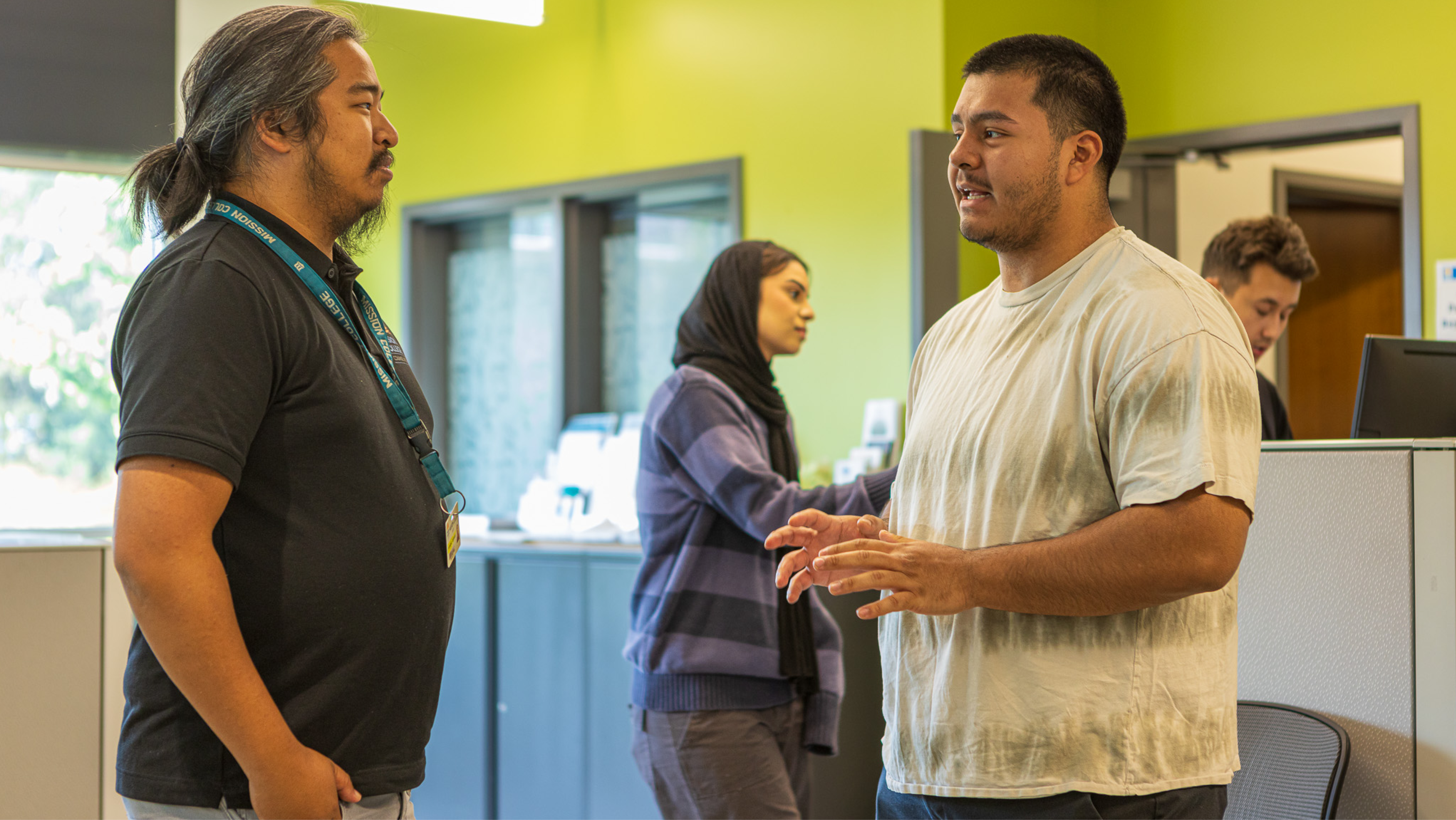 A male staff member in a Mission College lanyard speaking with a male student in the counseling area.