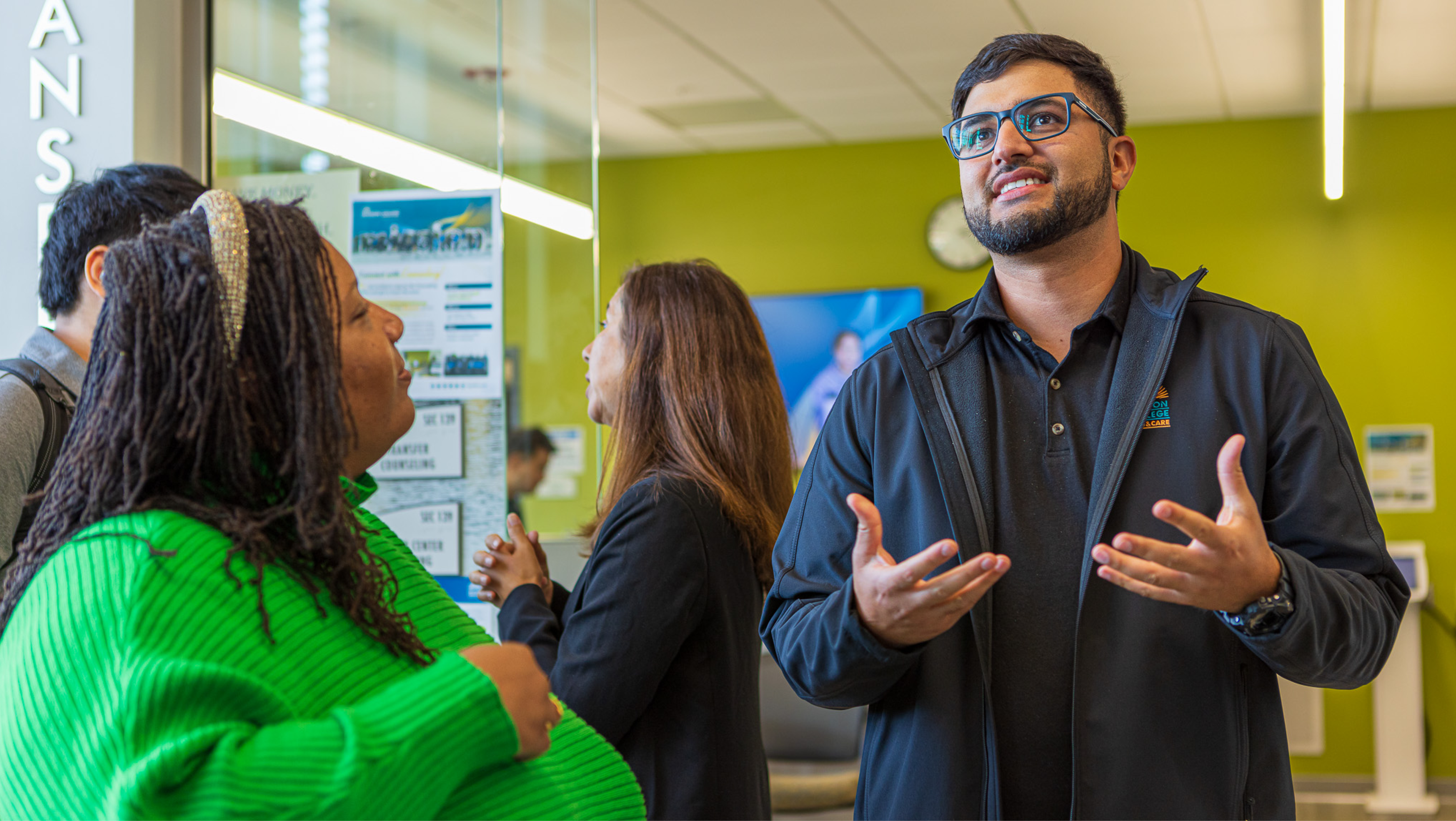 A male Mission College staff member in a black jacket gesturing while speaking with a student in the lobby.