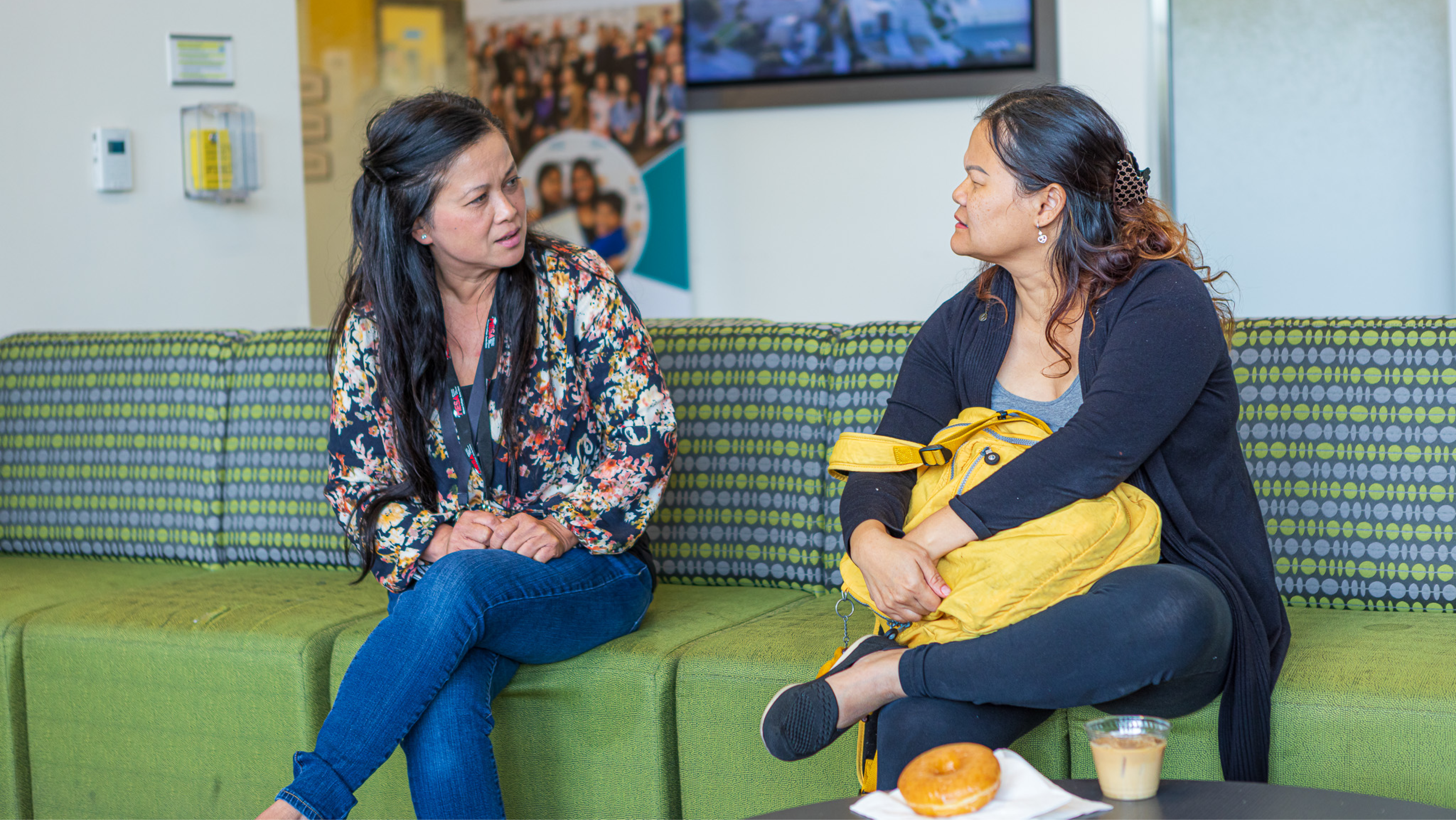 Two women sitting and chatting on a green couch in a student lounge area near a coffee table.