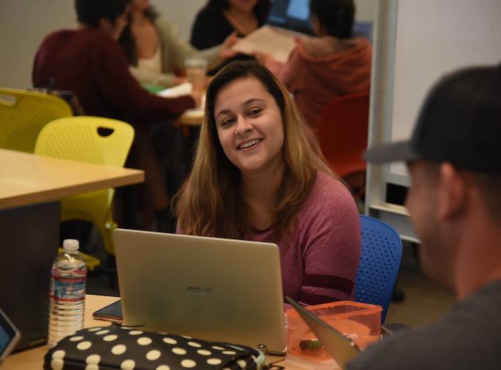 Young woman with long black hair works at a laptop in the library. She sits at a table with other students.