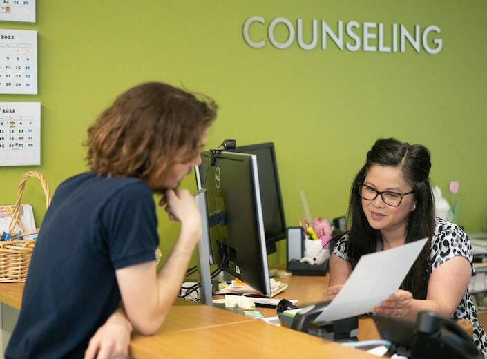 Counselor helping student in Counseling Center. She has shoulder-length black hair and glasses. She points to a paper as she assists a young man with curly brown hair.