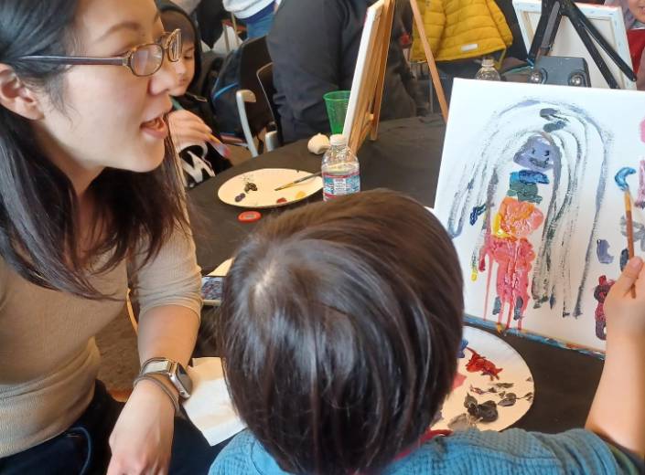 Young Asian woman with glasses and straight black hair works with a young boy around four as he works on a painting.
