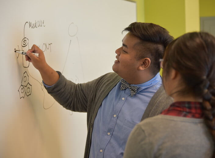 Two students work together at a markerboard. Both are of Asian descent. The male student wears a light blue shirt and bowtie, he draws on the board. The young woman stands near him and observes/listens.