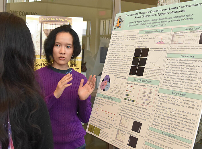 Asian-American female student in purple sweater explains her project to someone at an event.