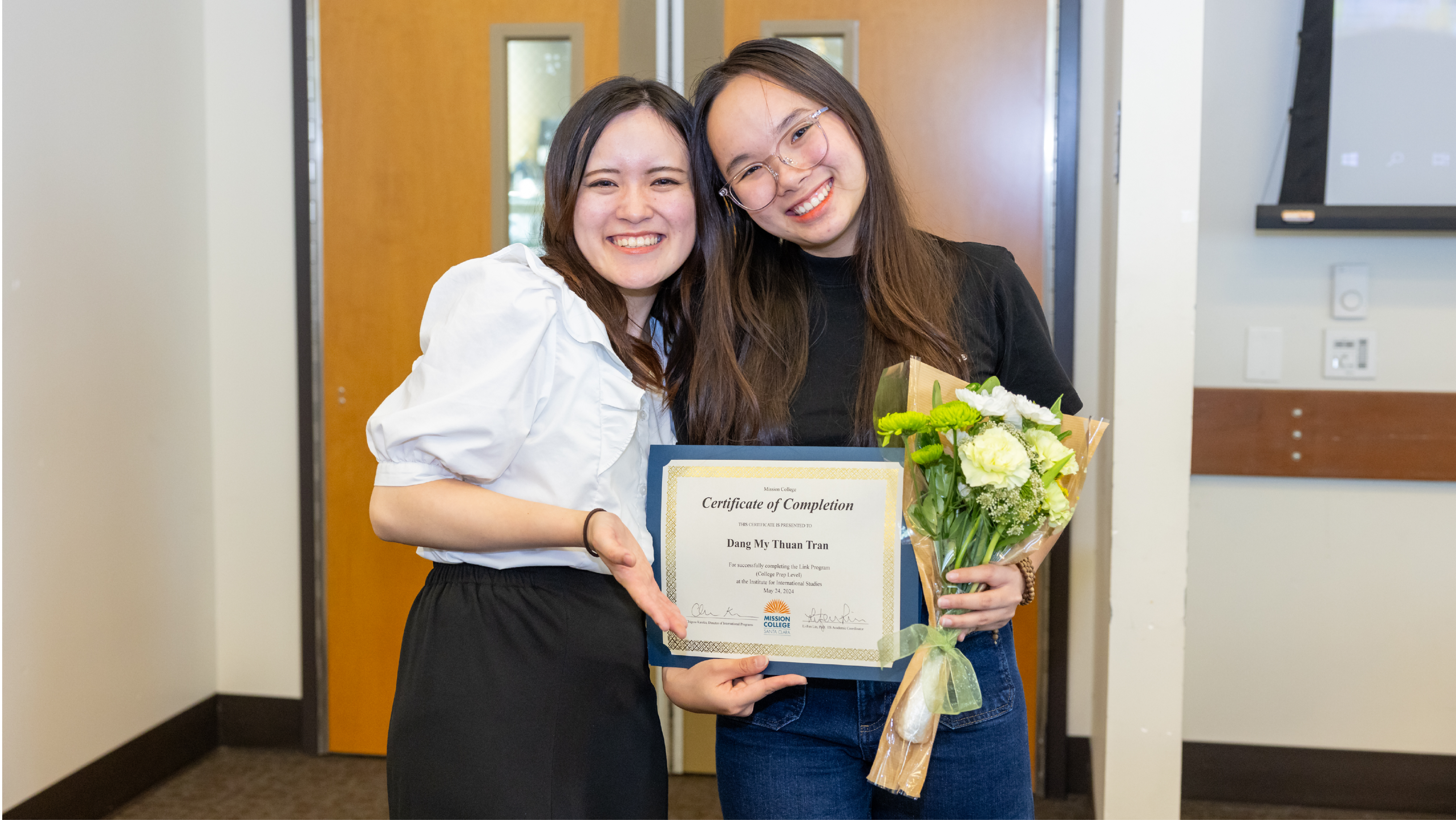 Two students smiling as one presents a framed Certificate of Completion and a bouquet of flowers to the other.