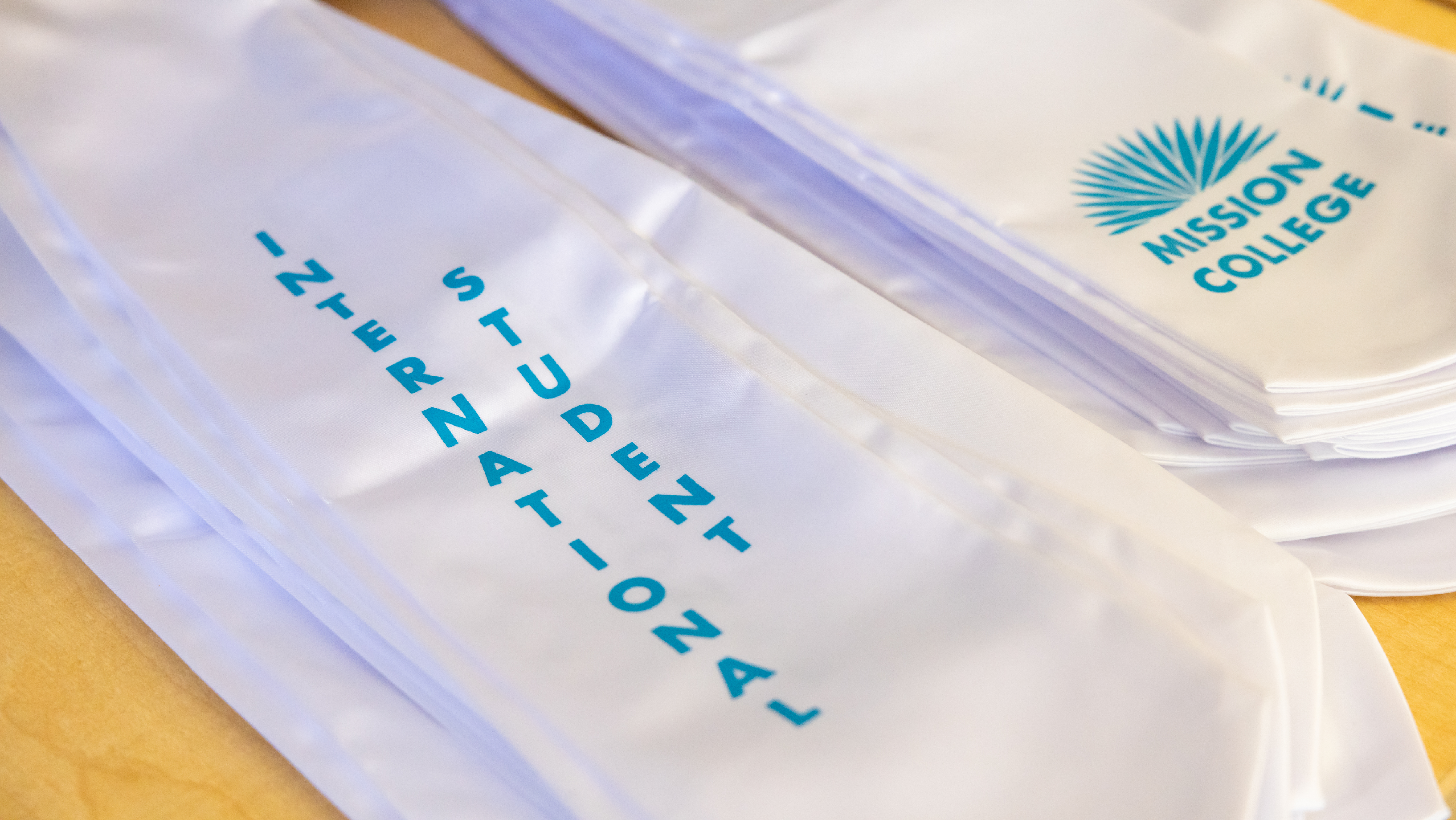 Close-up of white graduation sashes printed with "International Student" and the Mission College logo stacked on a table.