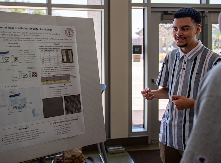 Student presents his research at an event. He is Latino and wears a striped blue and white shirt.