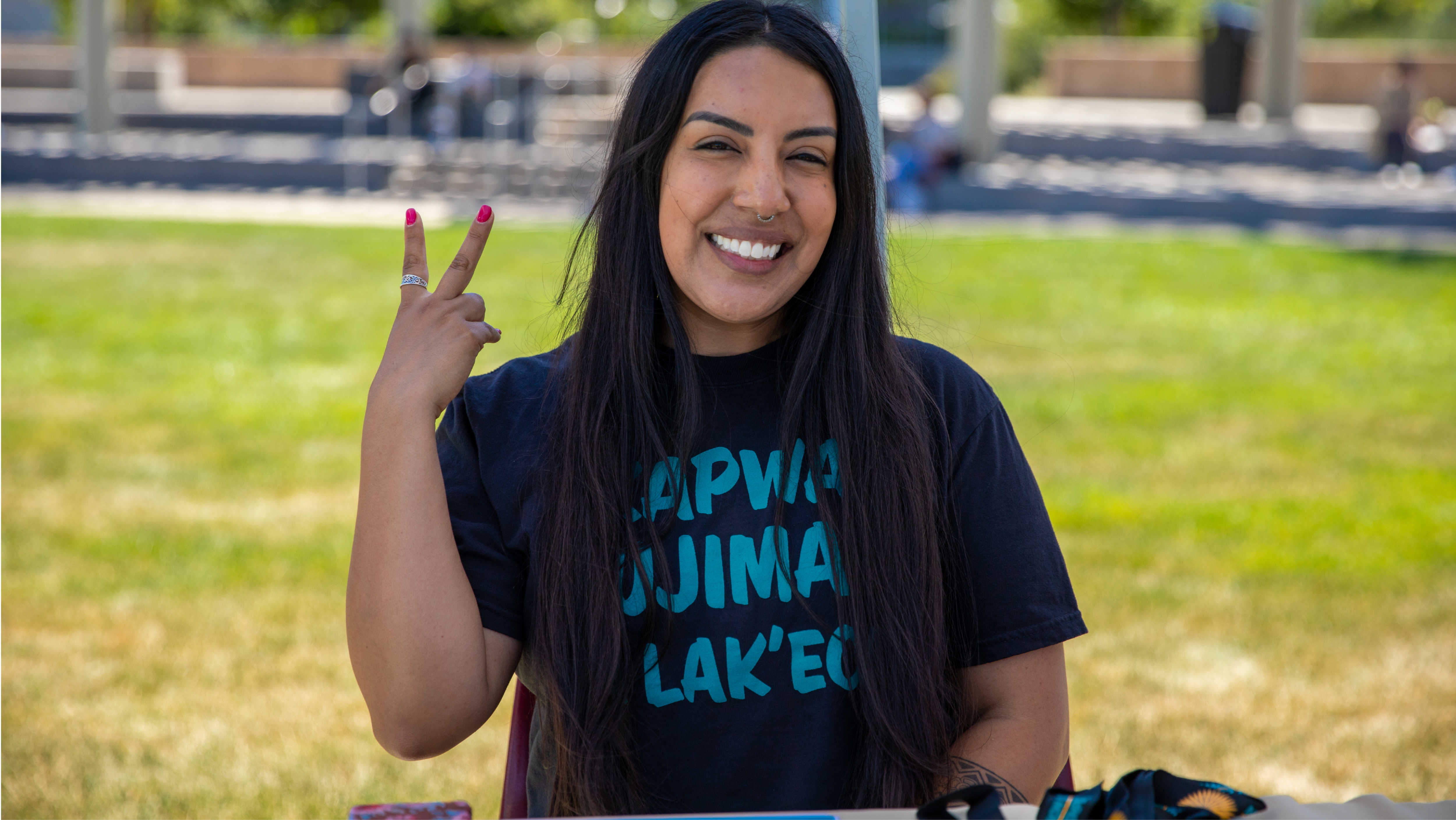 A smiling student wearing a black t-shirt giving a peace sign while sitting outdoors on campus.