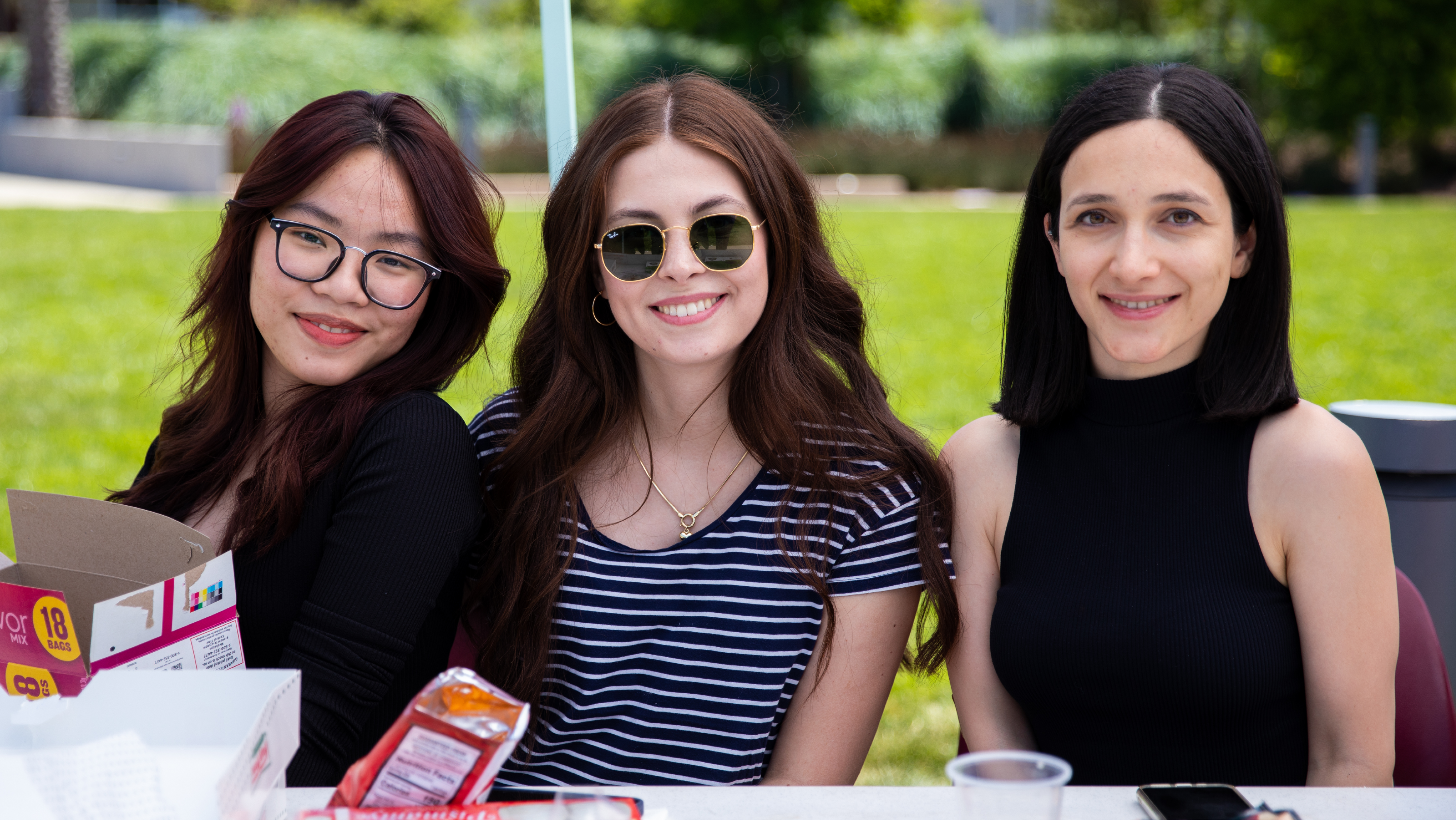 Three smiling female students seated together outdoors at a campus event table.