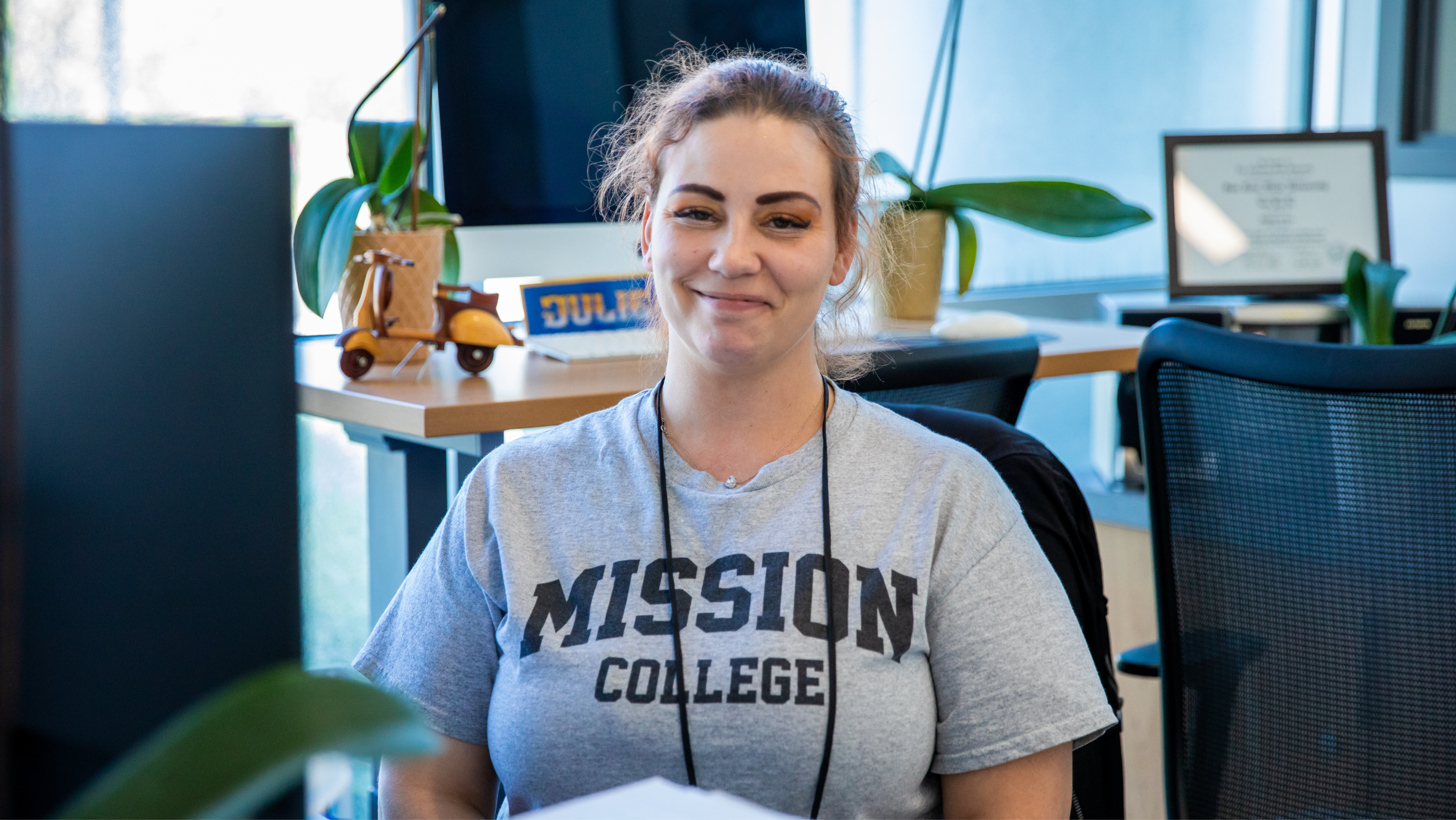 A smiling Mission College staff member sitting at an office desk wearing a grey heather Mission College shirt.