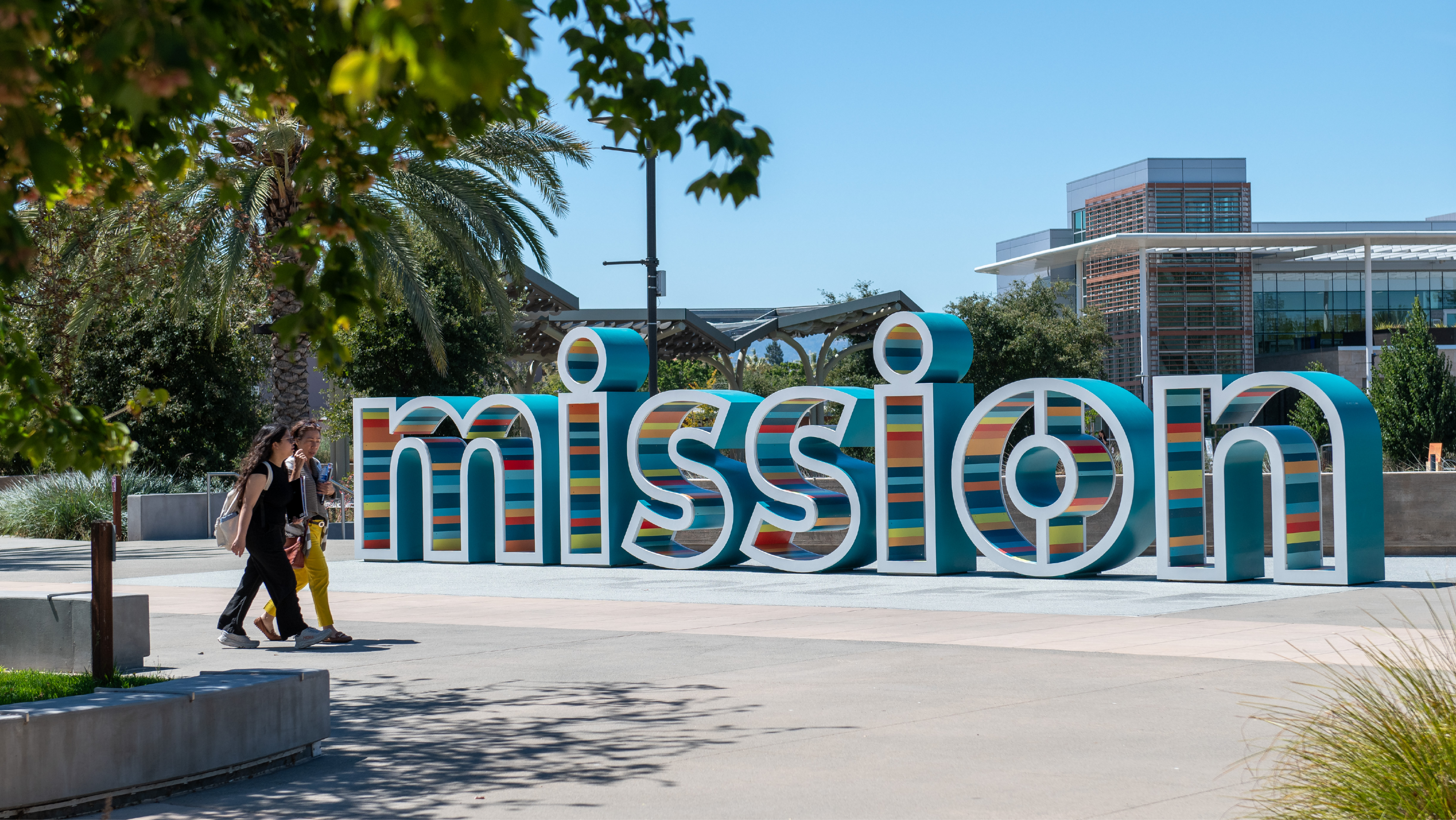 Two people walking past the large colorful Mission letter sign sculpture in the central campus plaza.