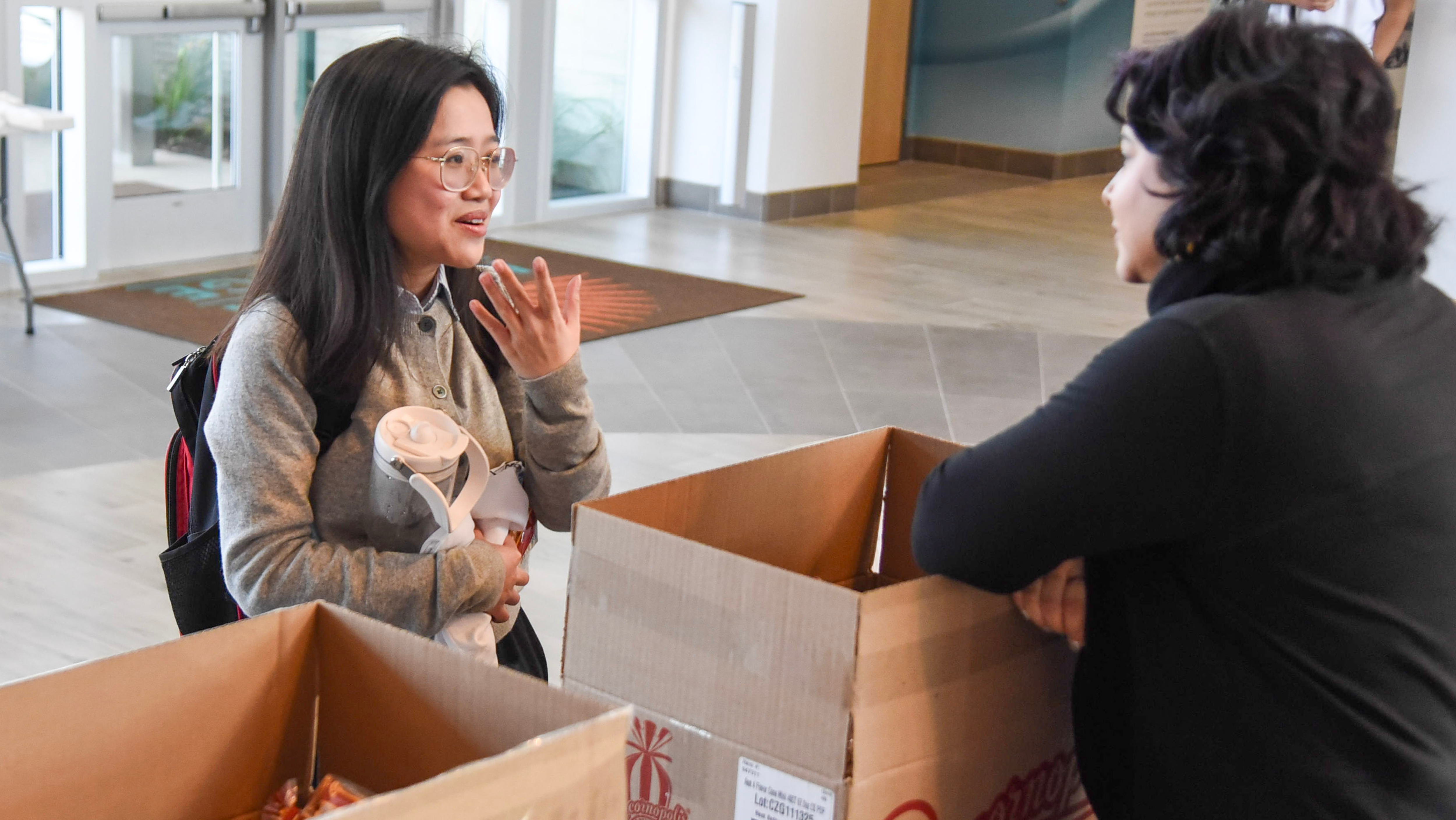 A student holding a water bottle talking with a staff member at an indoor event check-in table.