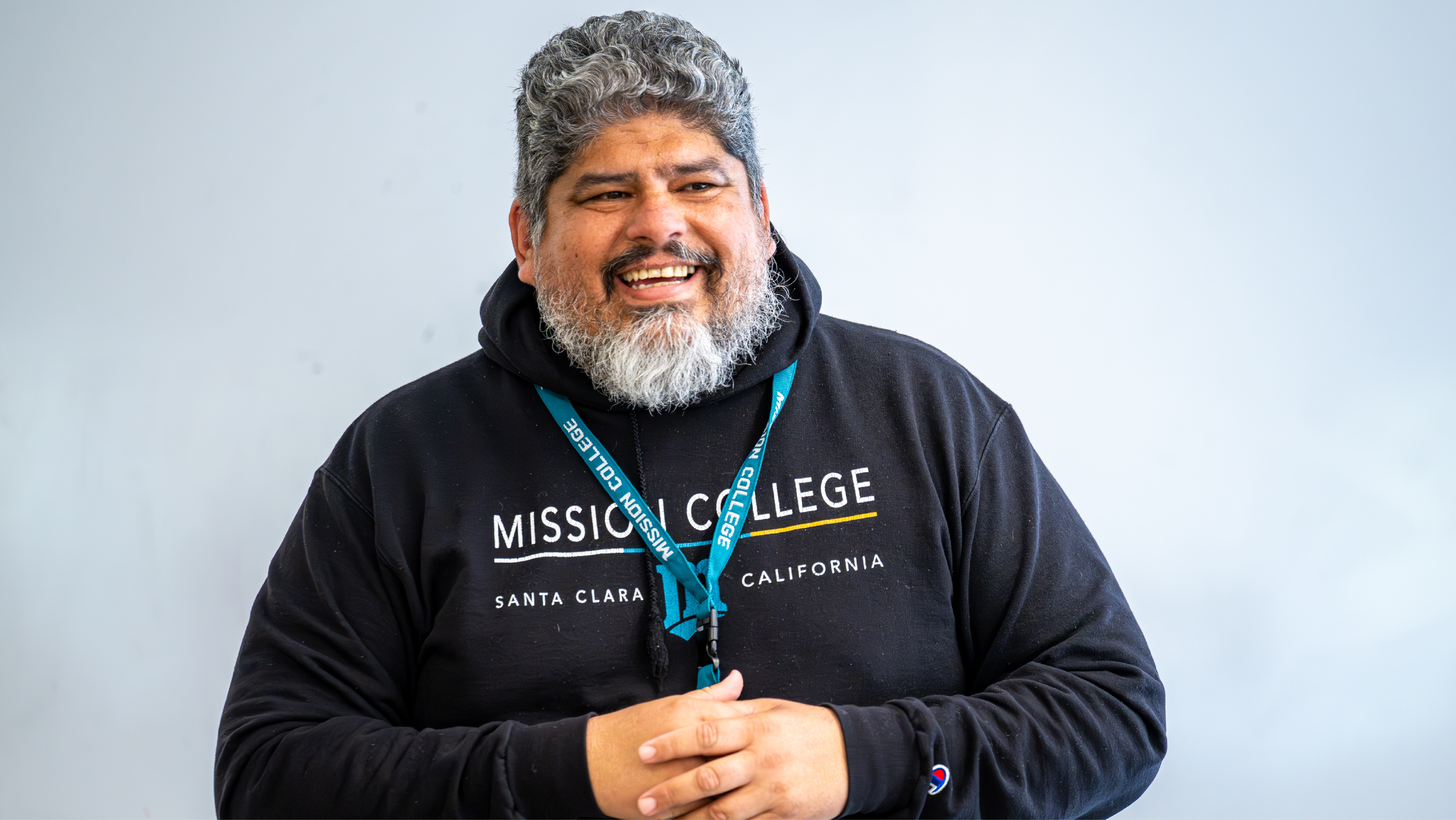 A smiling staff member wearing a black Mission College hoodie and lanyard clasps his hands during a conversation.