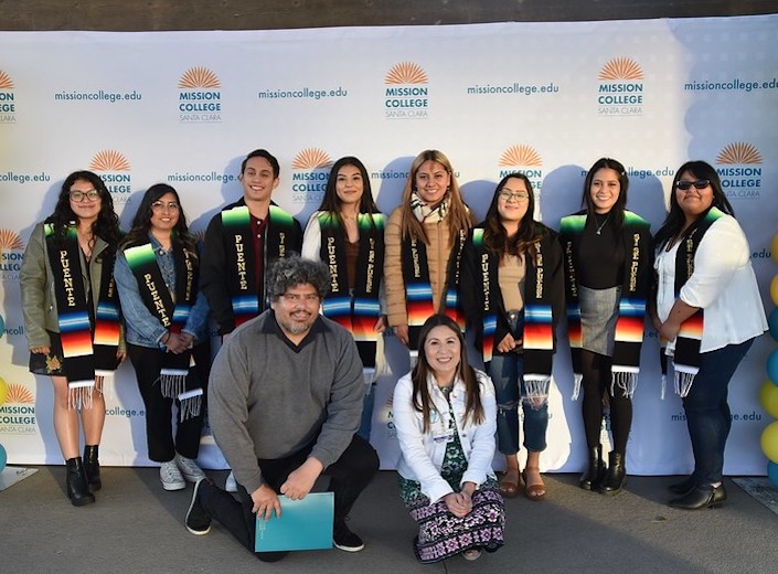 Group of Puente students pose in front of a "step and repeat" backdrop wearing colorful woven Puente sashes.