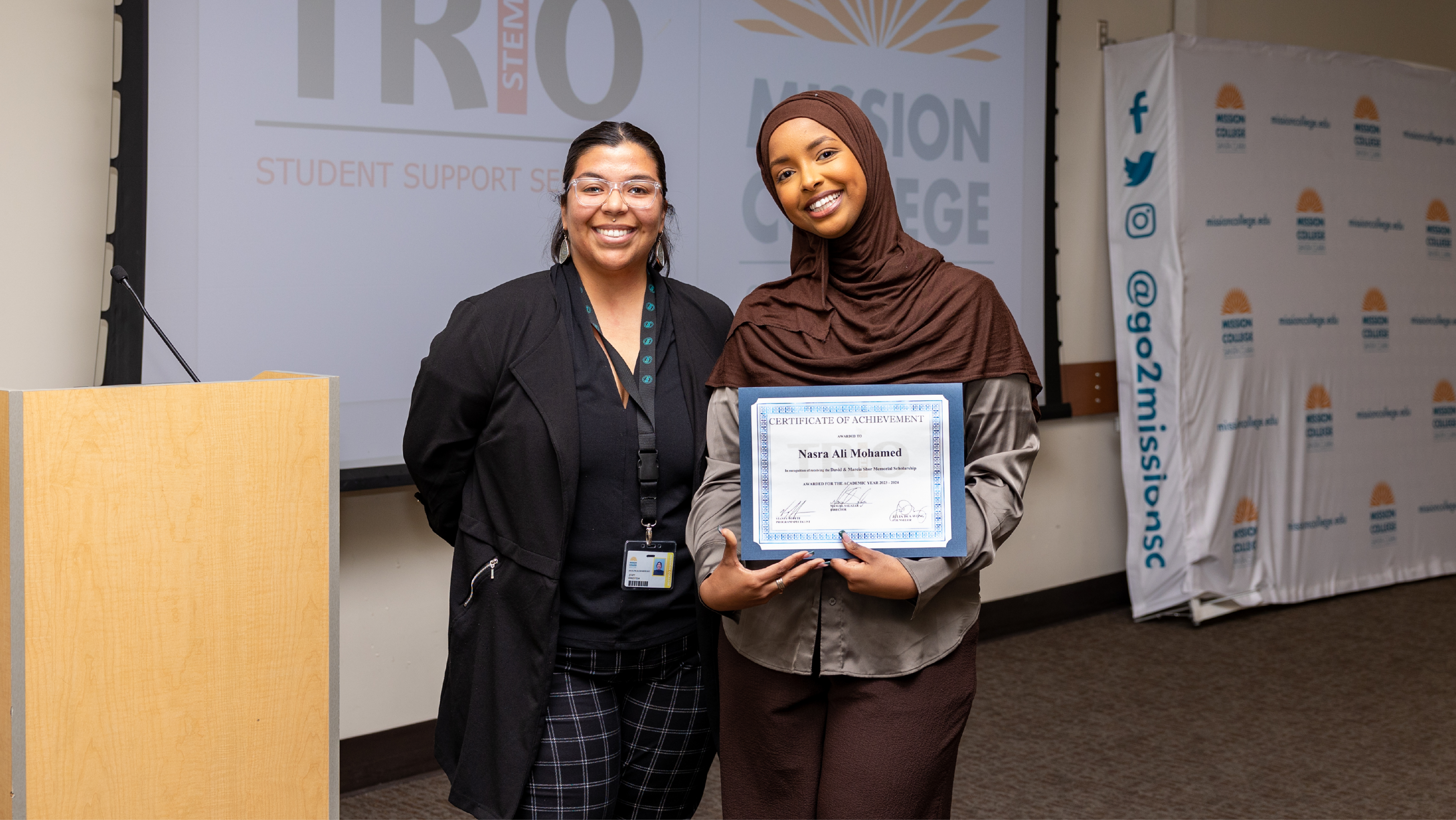 A student holding a framed certificate of achievement poses with a staff member in front of a TRIO Student Support Services backdrop.