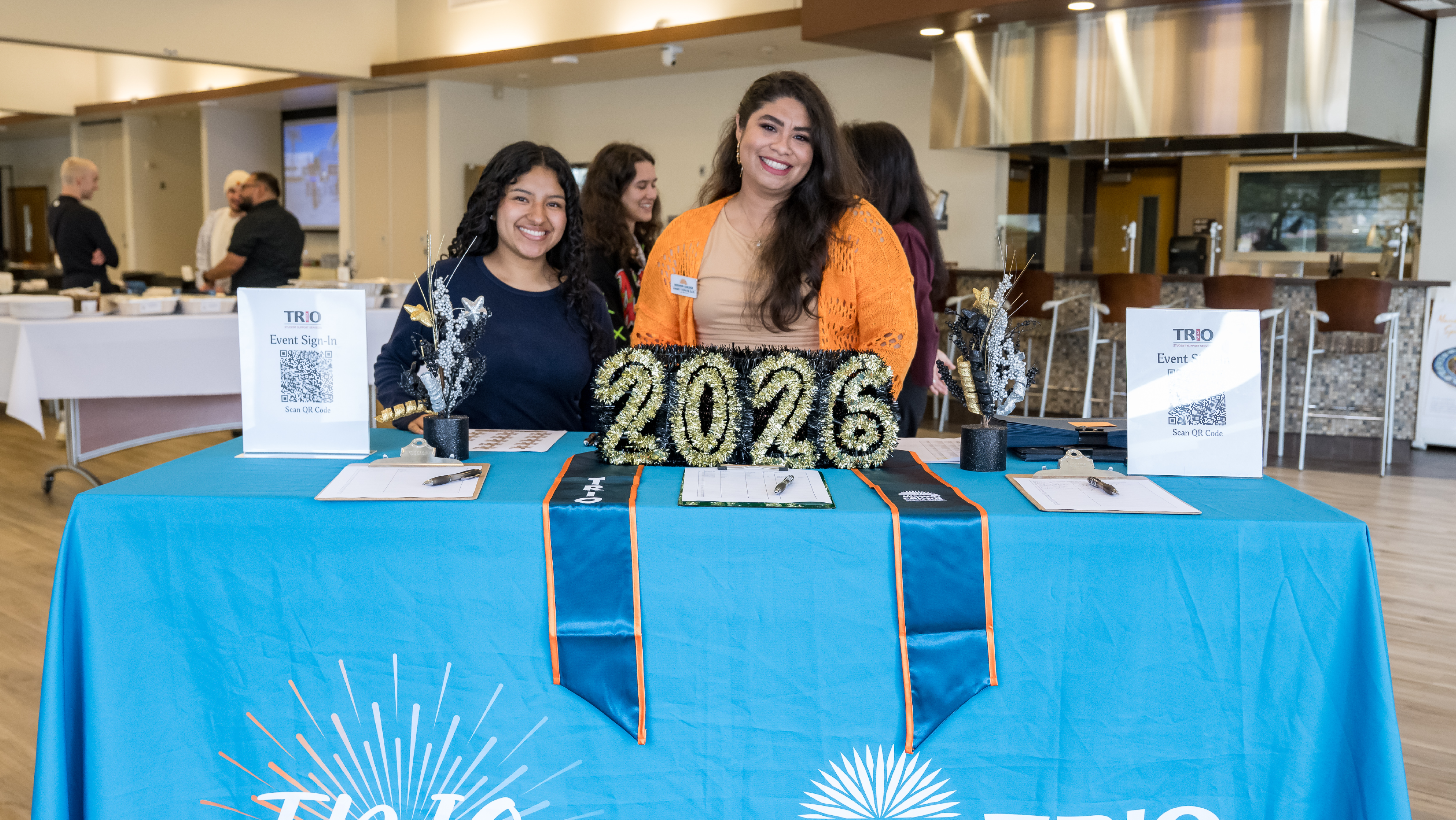 Two staff members smiling behind a blue check-in table decorated with 2026 numbers and TRIO graduation stoles at a campus event.