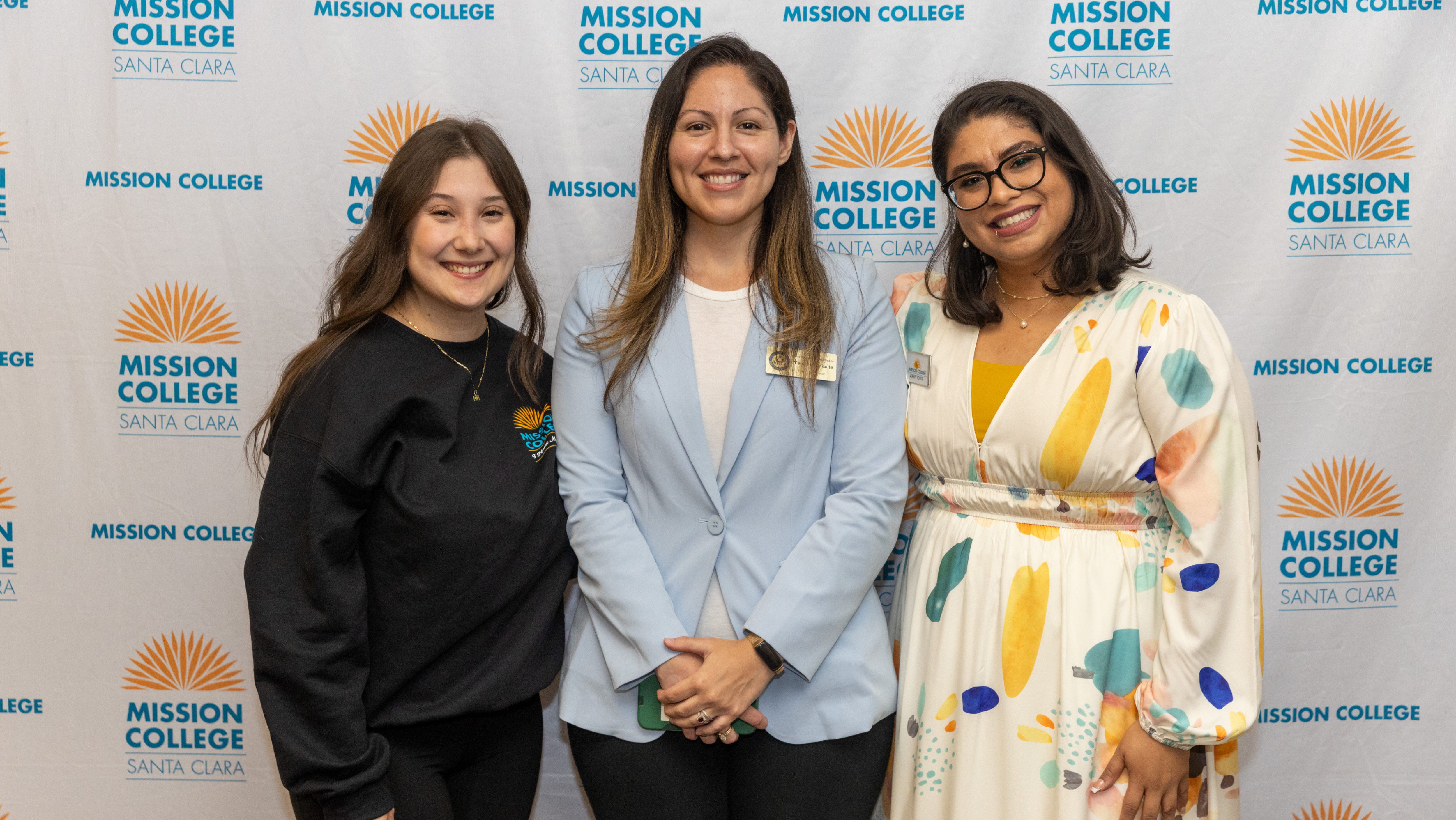 Three Mission College community members smiling together in front of a branded backdrop at a campus recognition event.