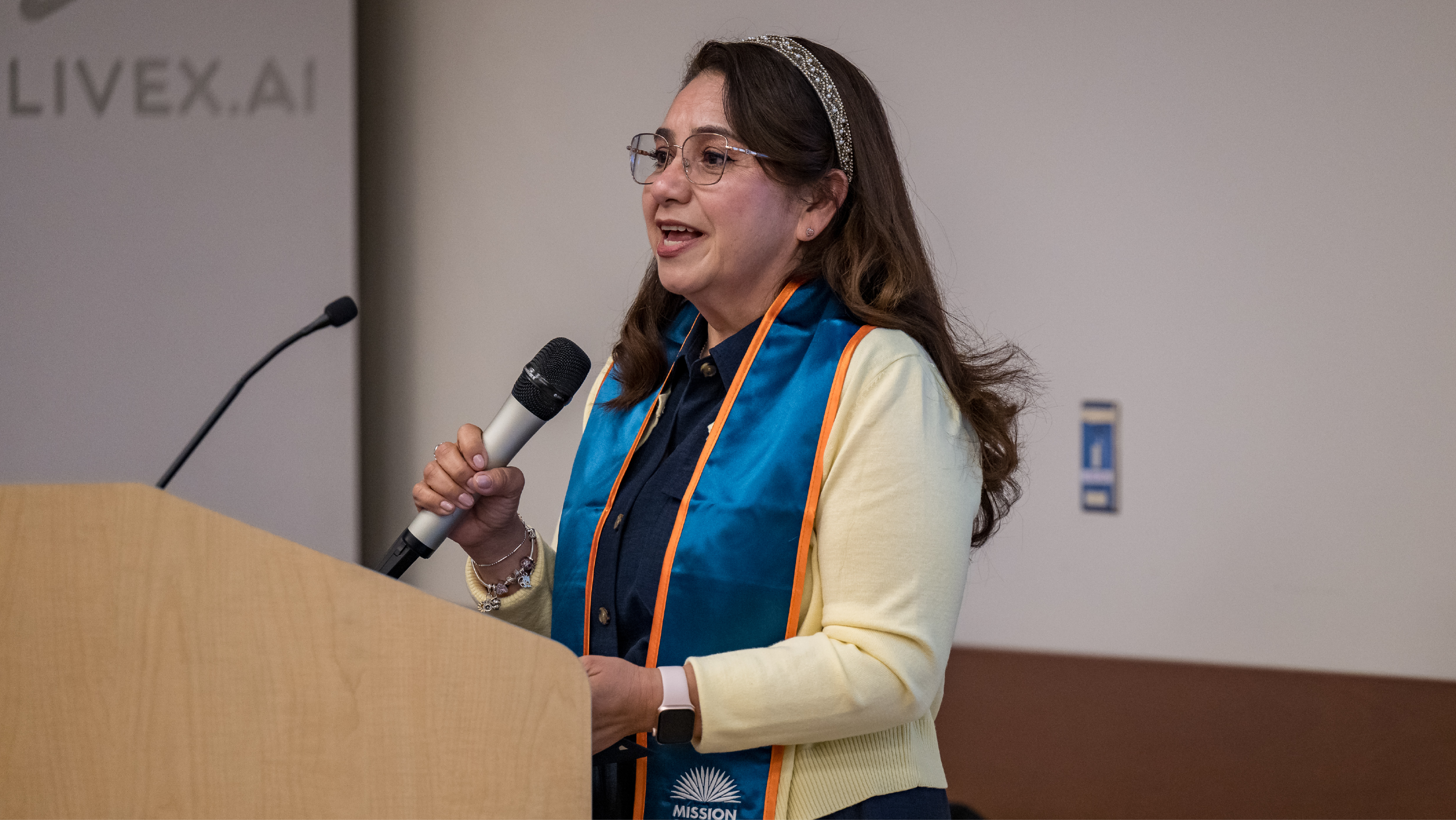 A female student wearing a teal TRIO graduation stole speaking into a microphone at a podium during a ceremony.