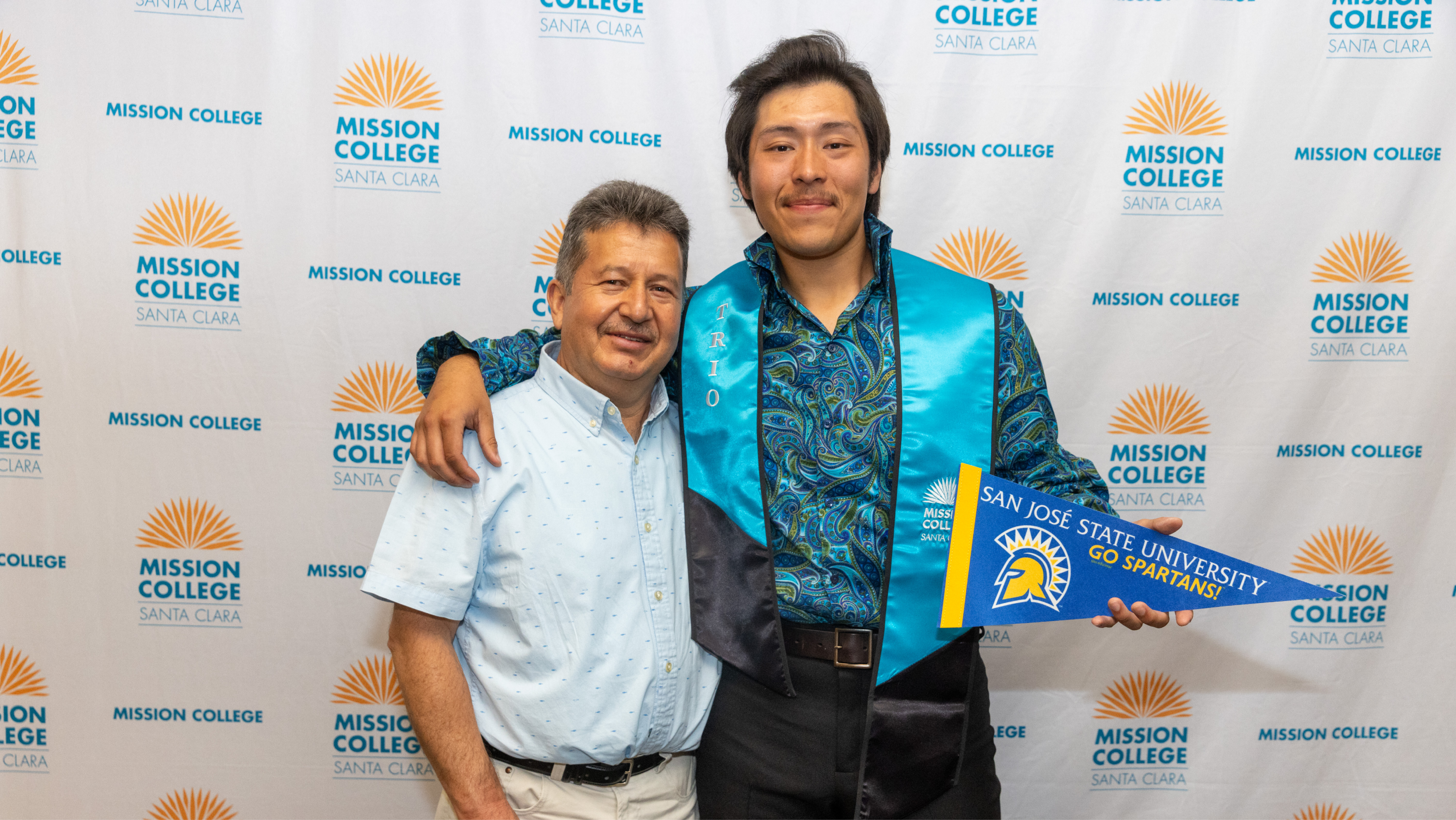 A graduating student in a TRIO stole holding a San Jose State University pennant posing arm-in-arm with a family member in front of a Mission College backdrop.