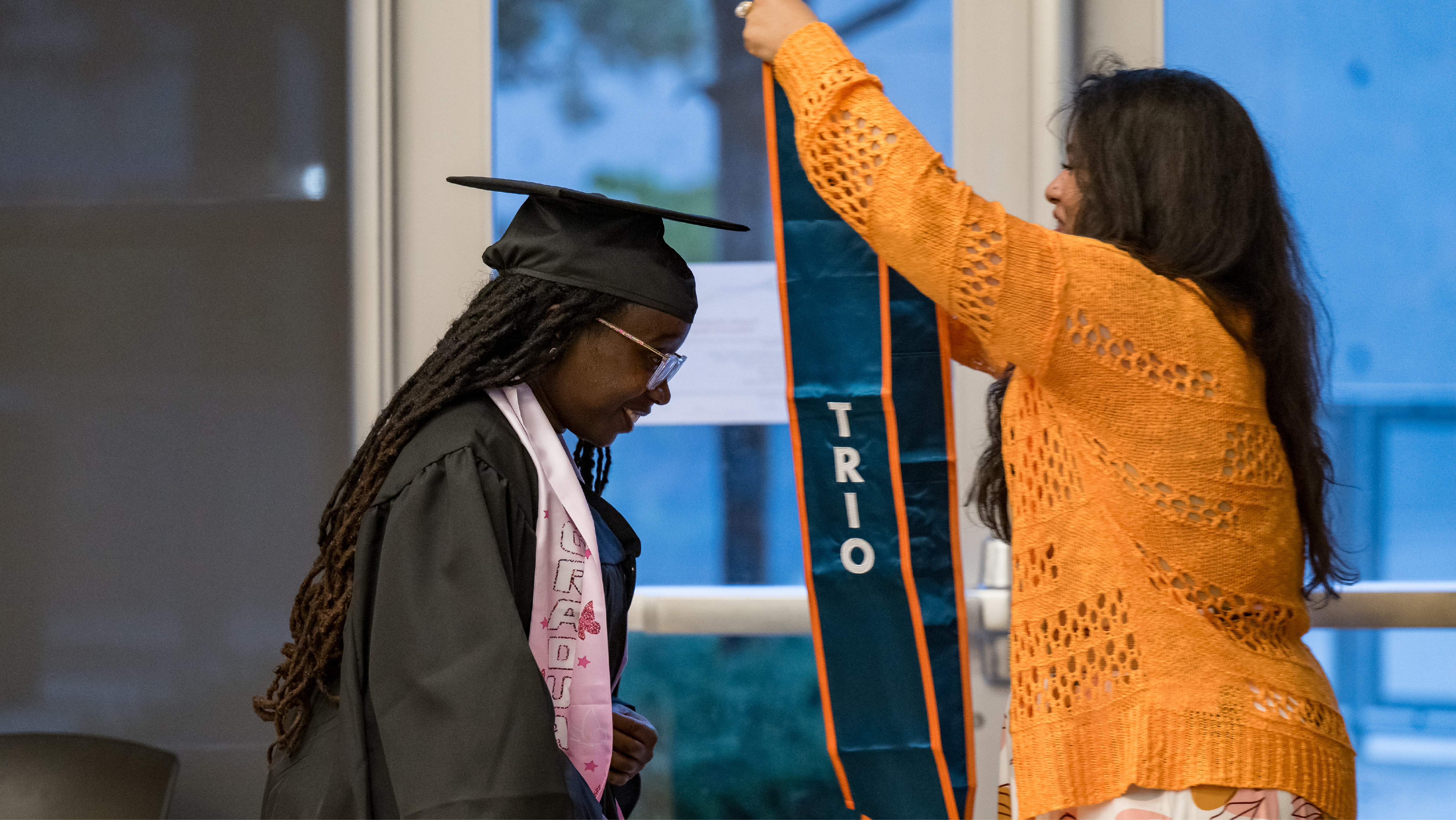 A staff member placing a teal TRIO graduation stole onto a student wearing a black cap and gown.