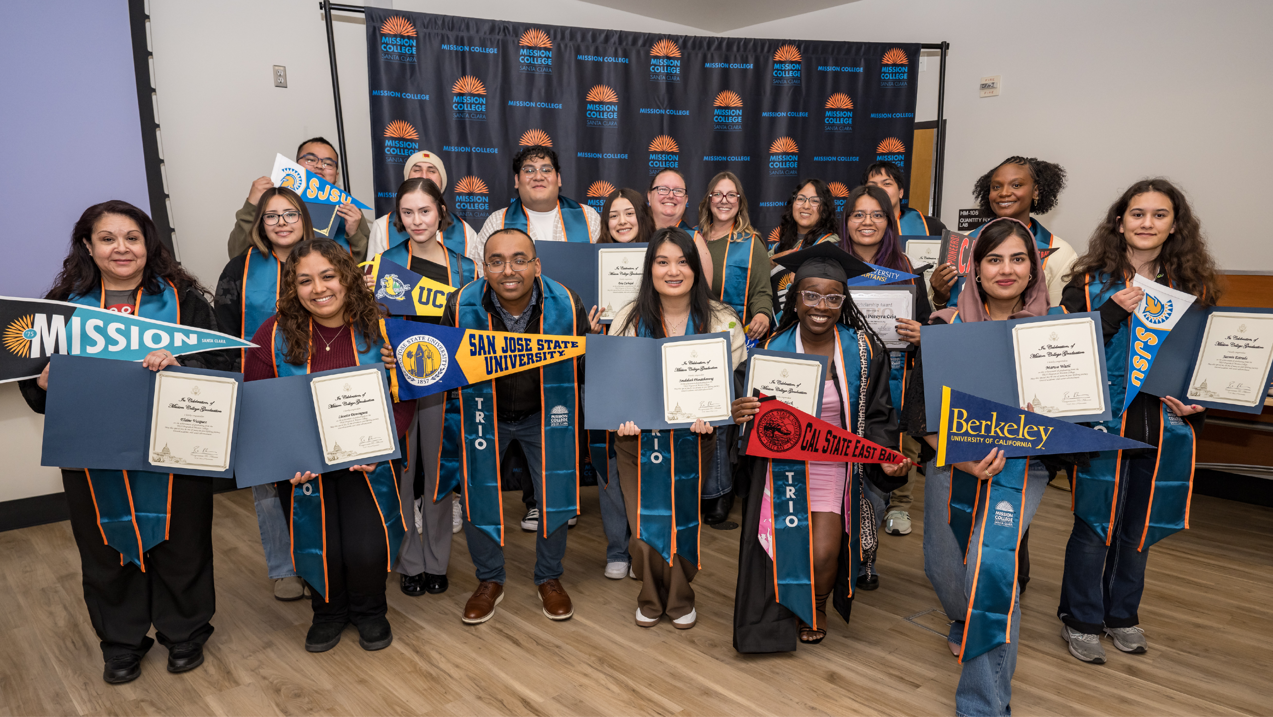 A large group of Mission College TRIO graduates and staff holding certificates, pennants, and signs in front of a backdrop at a celebration event.