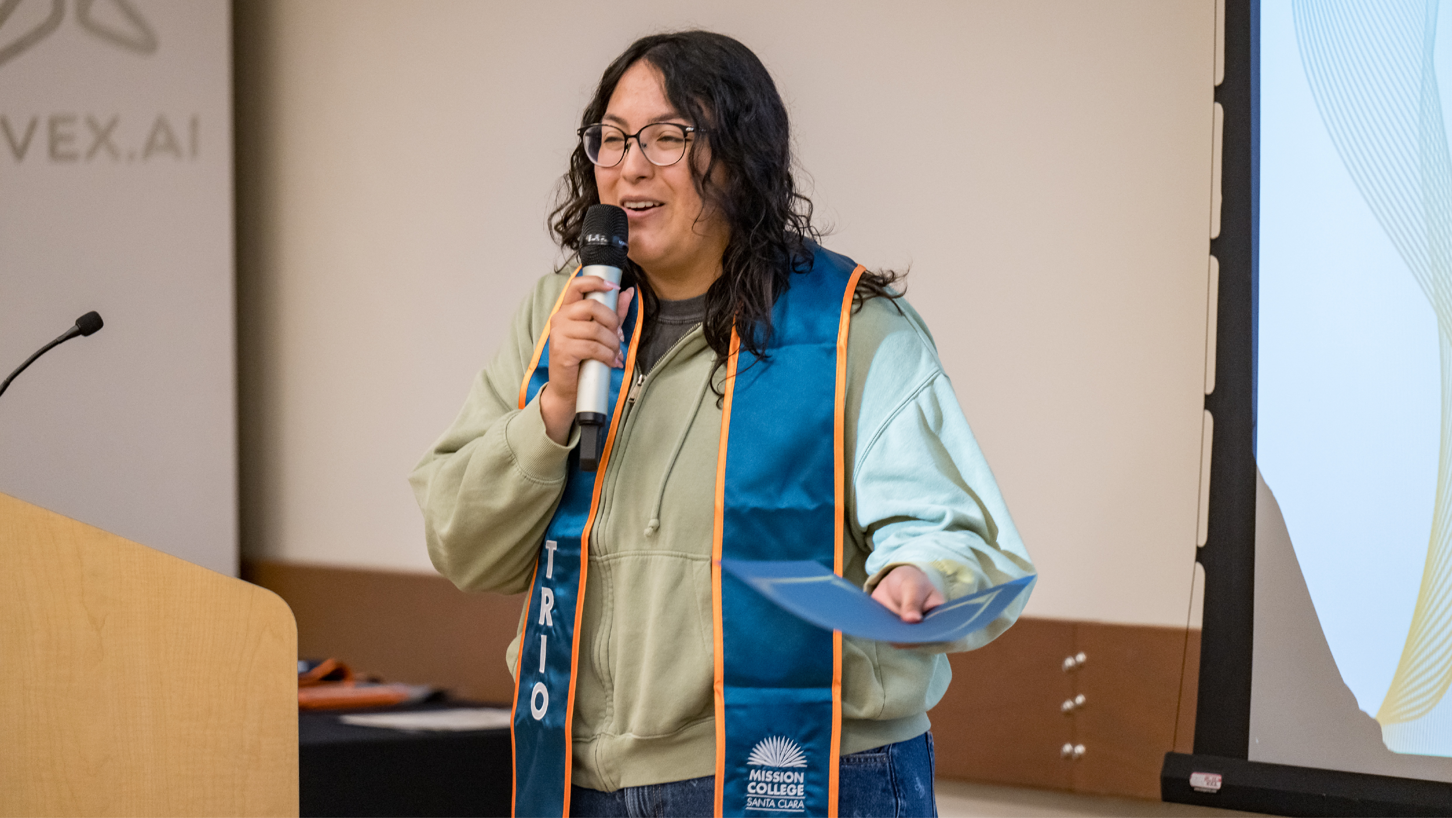 A student wearing a teal TRIO graduation stole speaking at a podium while holding a certificate folder.