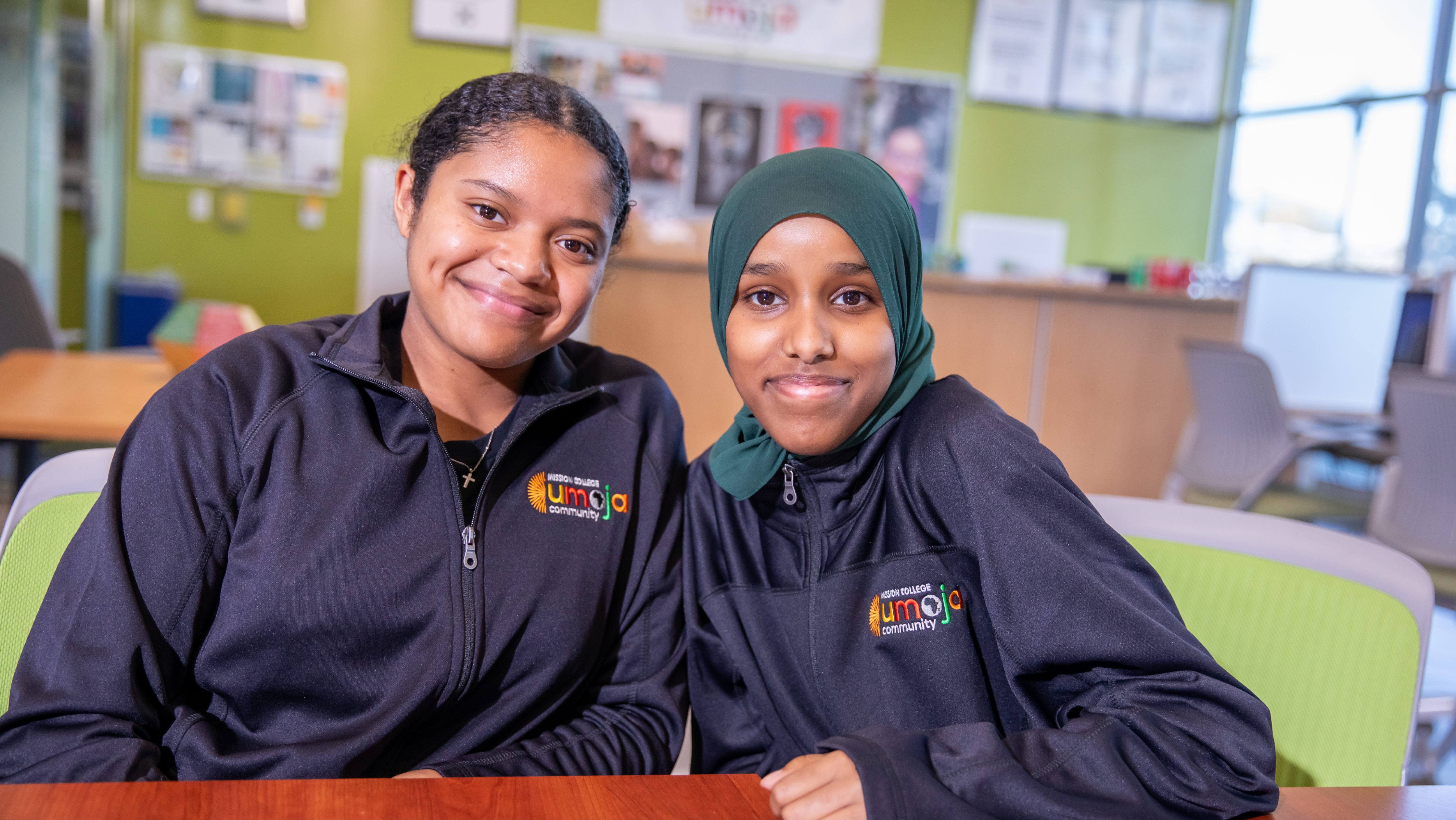 Two female students wearing matching Mission College Umoja black zip-up jackets sitting together and smiling.