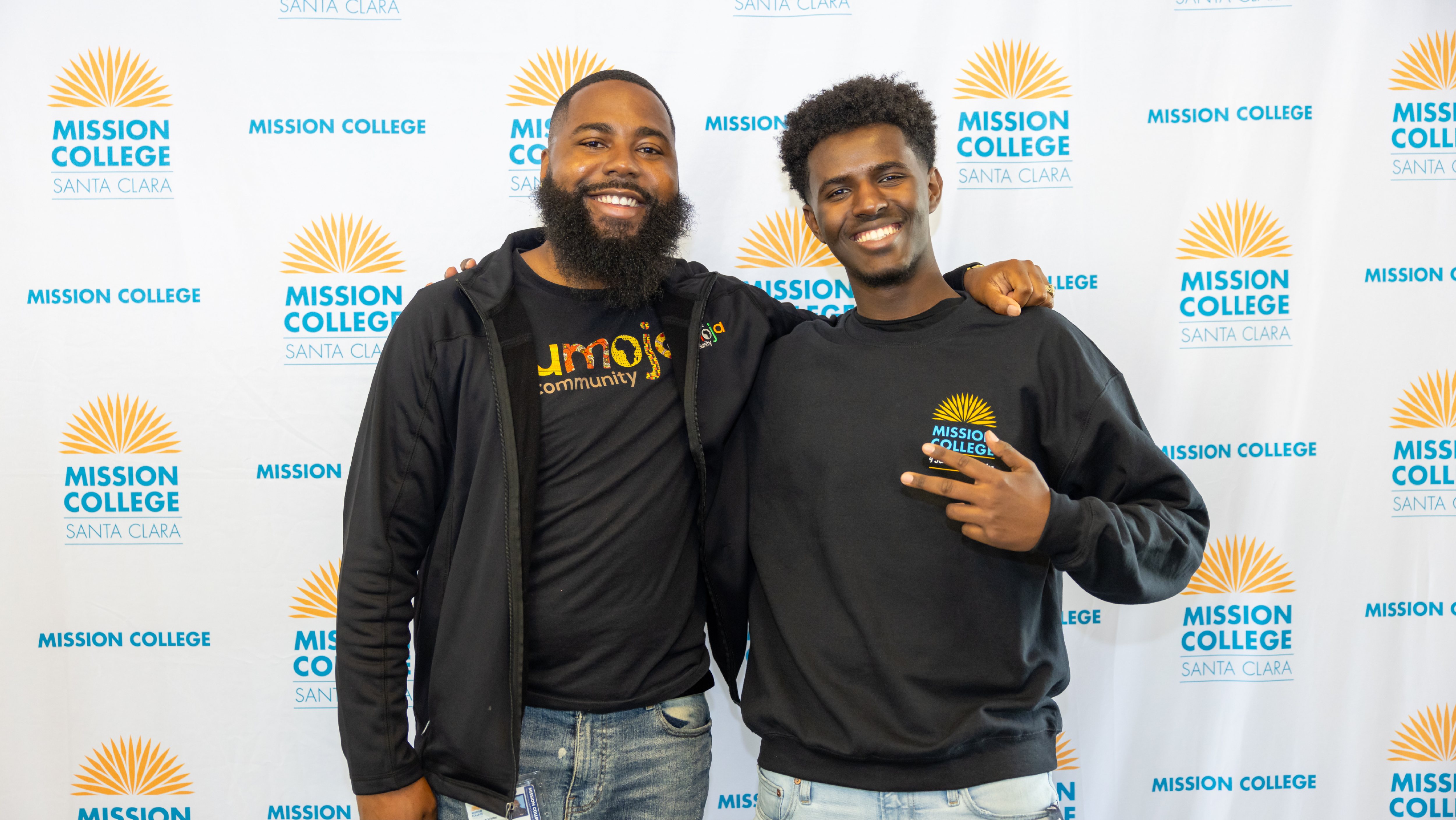 Two male students posing together with arms around each other in front of a branded Mission College backdrop.