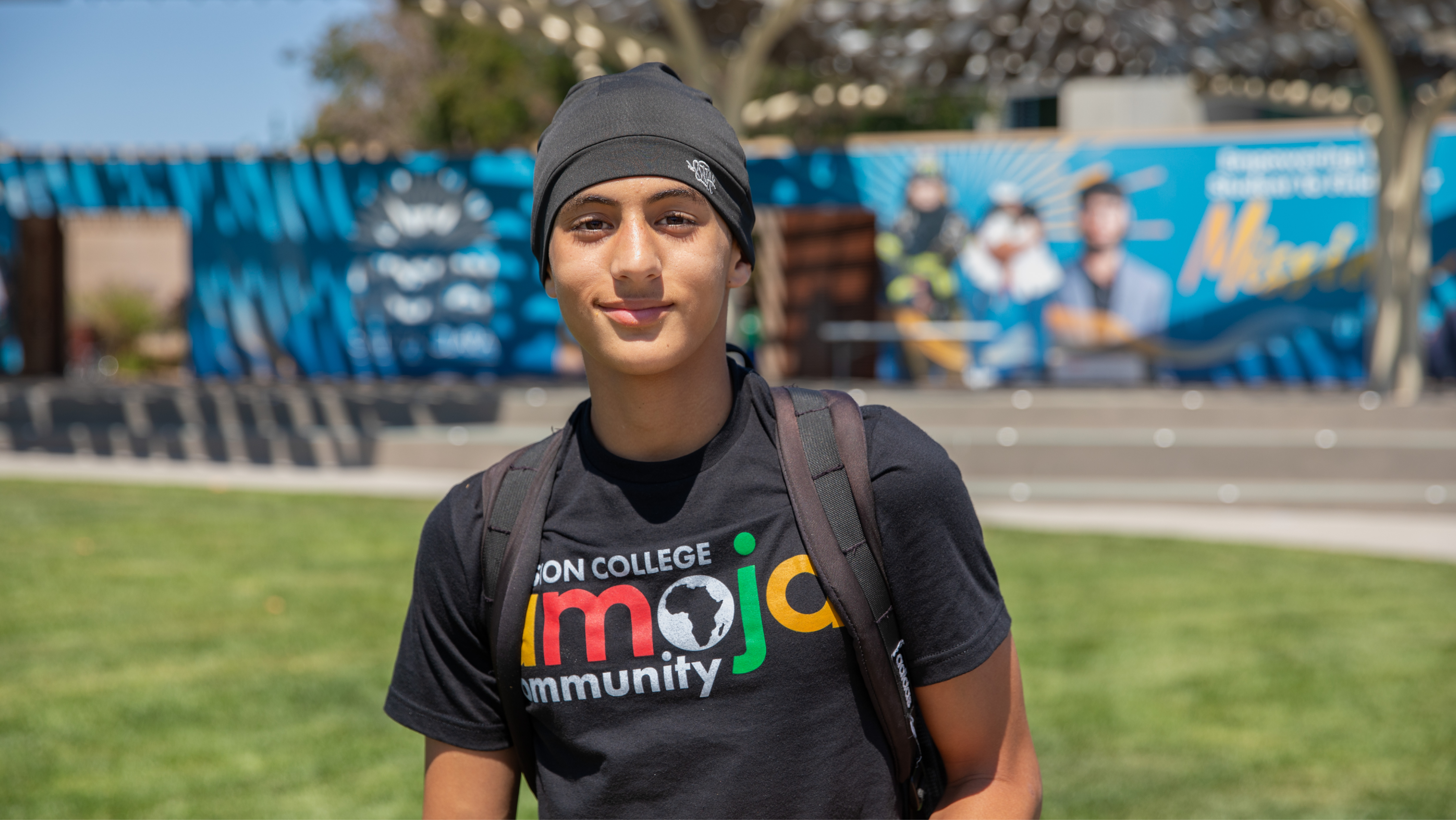 A student wearing a Mission College Umoja t-shirt and backpack smiling outdoors on campus.
