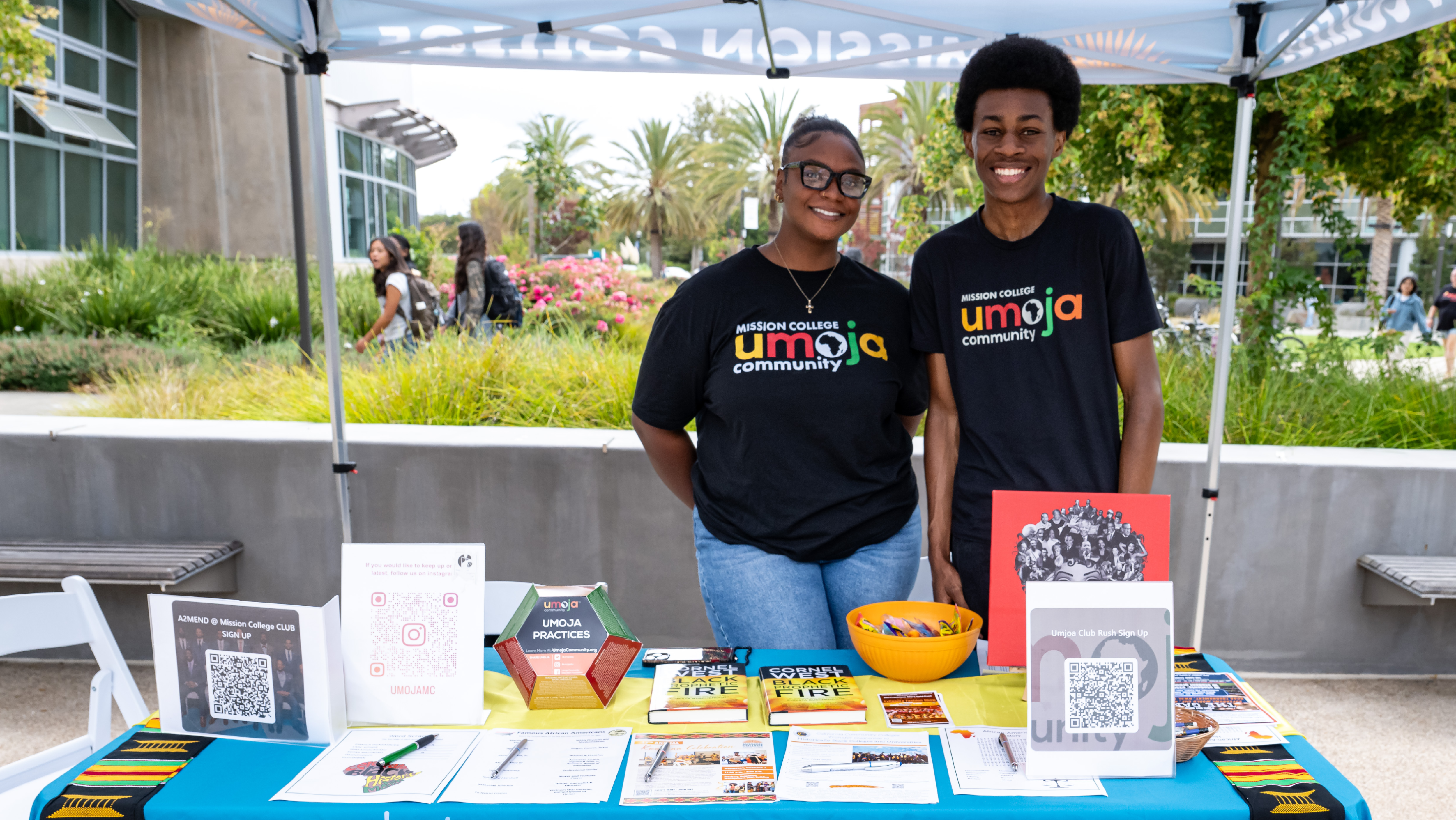 Two students wearing Mission College Umoja shirts smiling behind an information table with flyers, QR codes, and books under an outdoor tent.