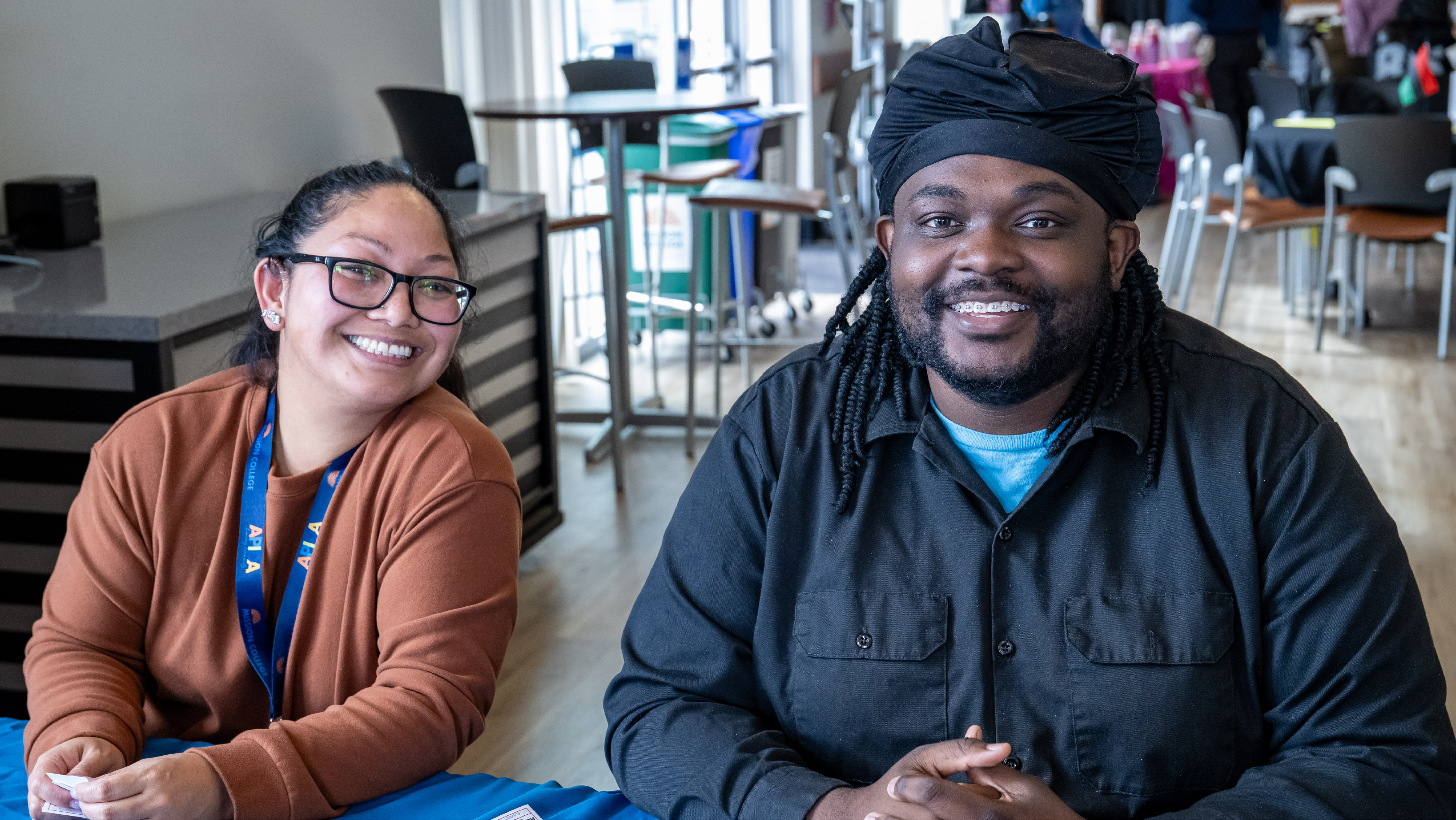 Two Mission College community members smiling together while seated at a campus event table.
