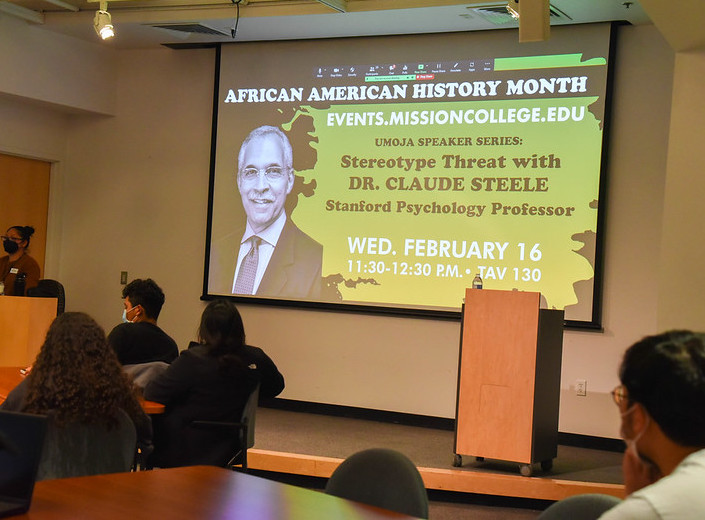 An event for Black History Month at Mission College. People are seated in a classroom at tables, a projecter shows a slide against a screen on the wall that reads "African American History Month. Umoja Spaker Series: Steregotype Thread with Dr, Claude Steel. Stanford Psychology Professor. Wednesday February 16, 11:30-12:30 p.m. - TAV 130.