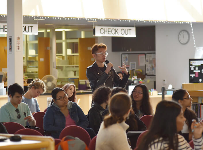 A young male student in a leather jackt and glasses holds a microphone as he asks a question at a literary event on Mission College campus.