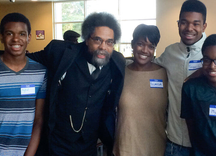 Dr. Cornel West event. He is a Black man in a loose suit with longish grey curly hair, glasses, and he has arms around a few students and faculty memebrs.