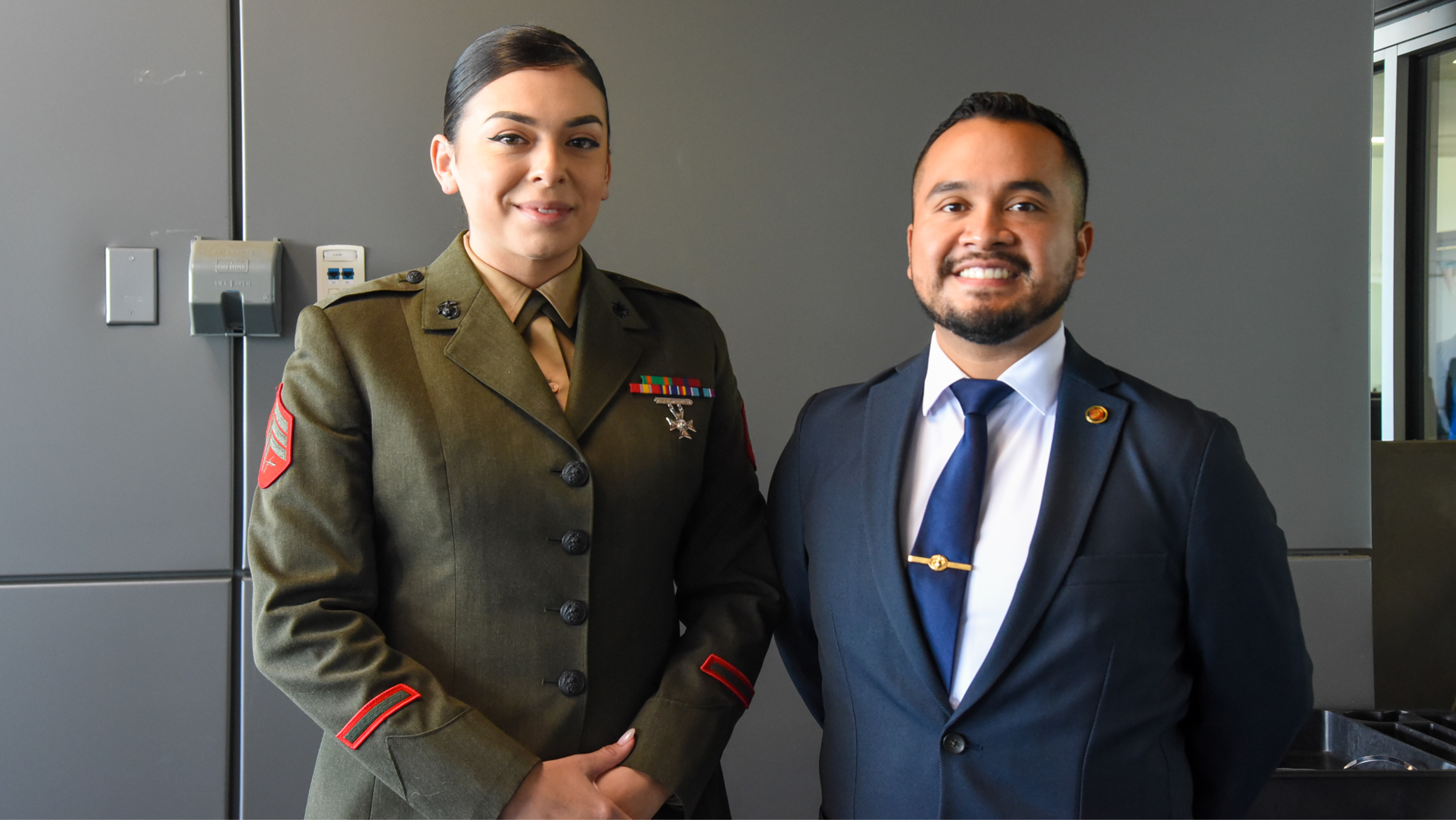 A female U.S. Marine in service uniform standing indoors next to a man in a dark suit and blue tie, both smiling for a photo.