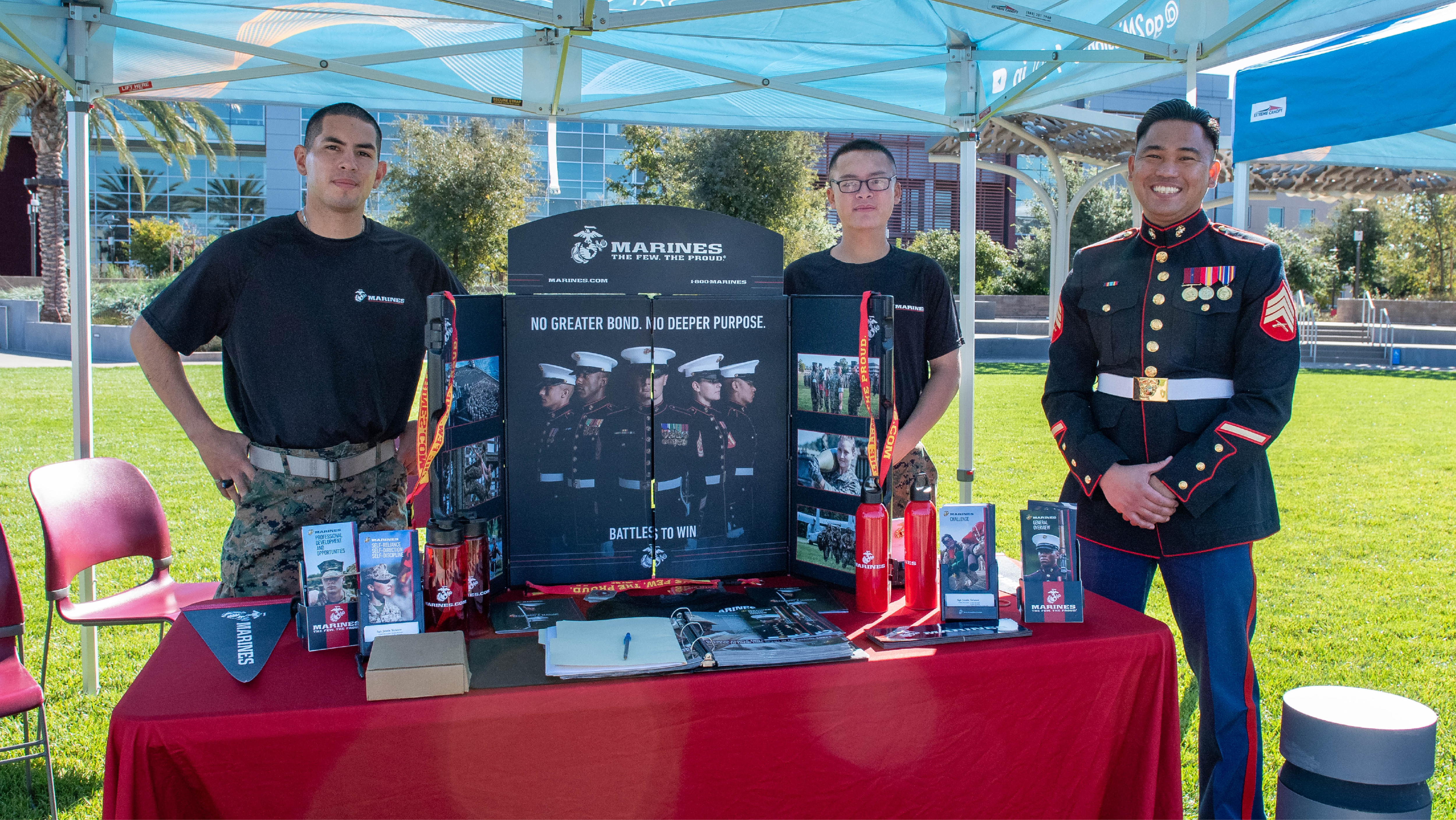 Two service members and a Marine Recruiter in full dress uniform posing at an outdoor U.S. Marine Corps information table.