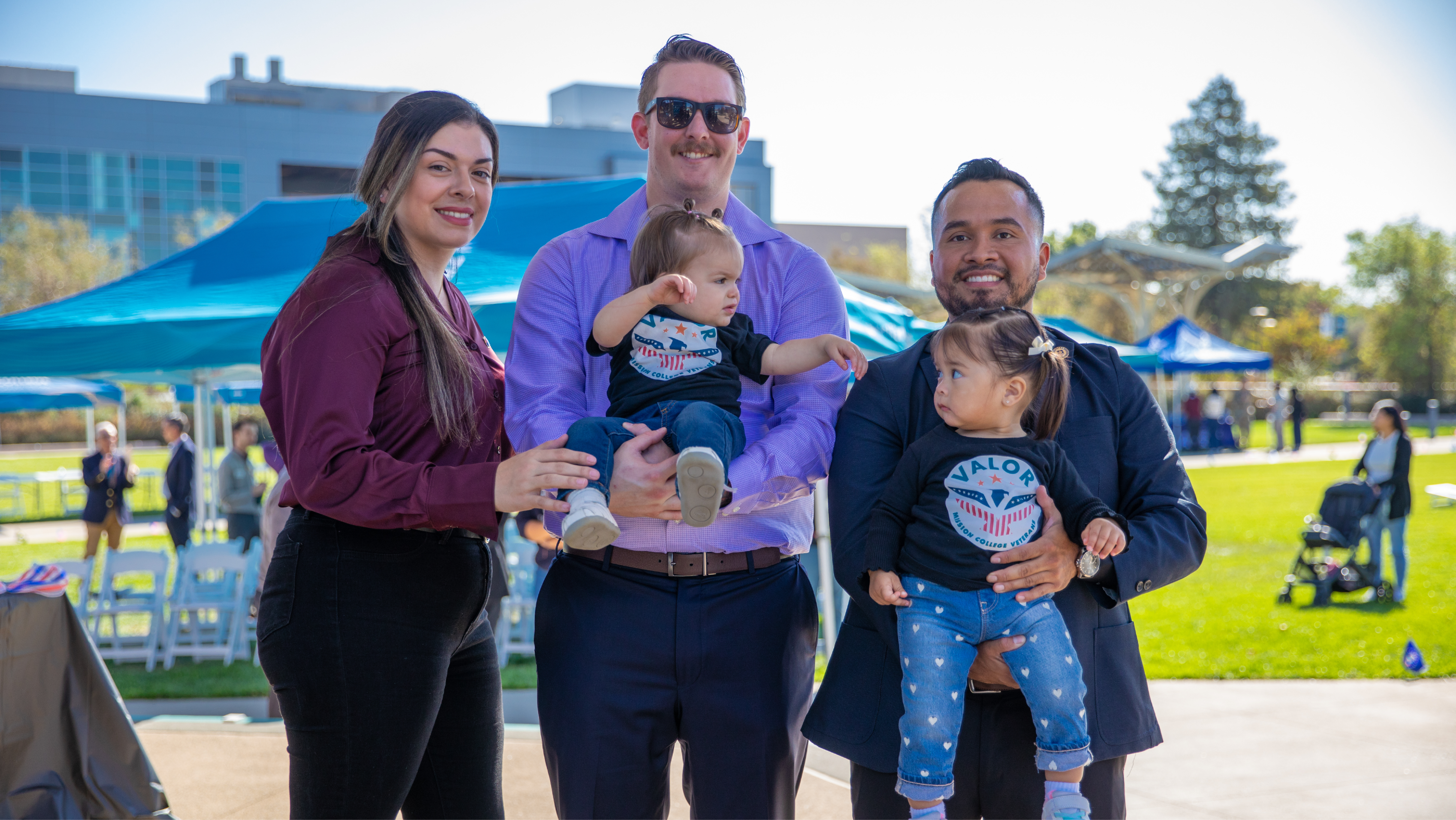 Two couples posing together outdoors on campus while holding two toddlers wearing Mission College VALOR shirts.