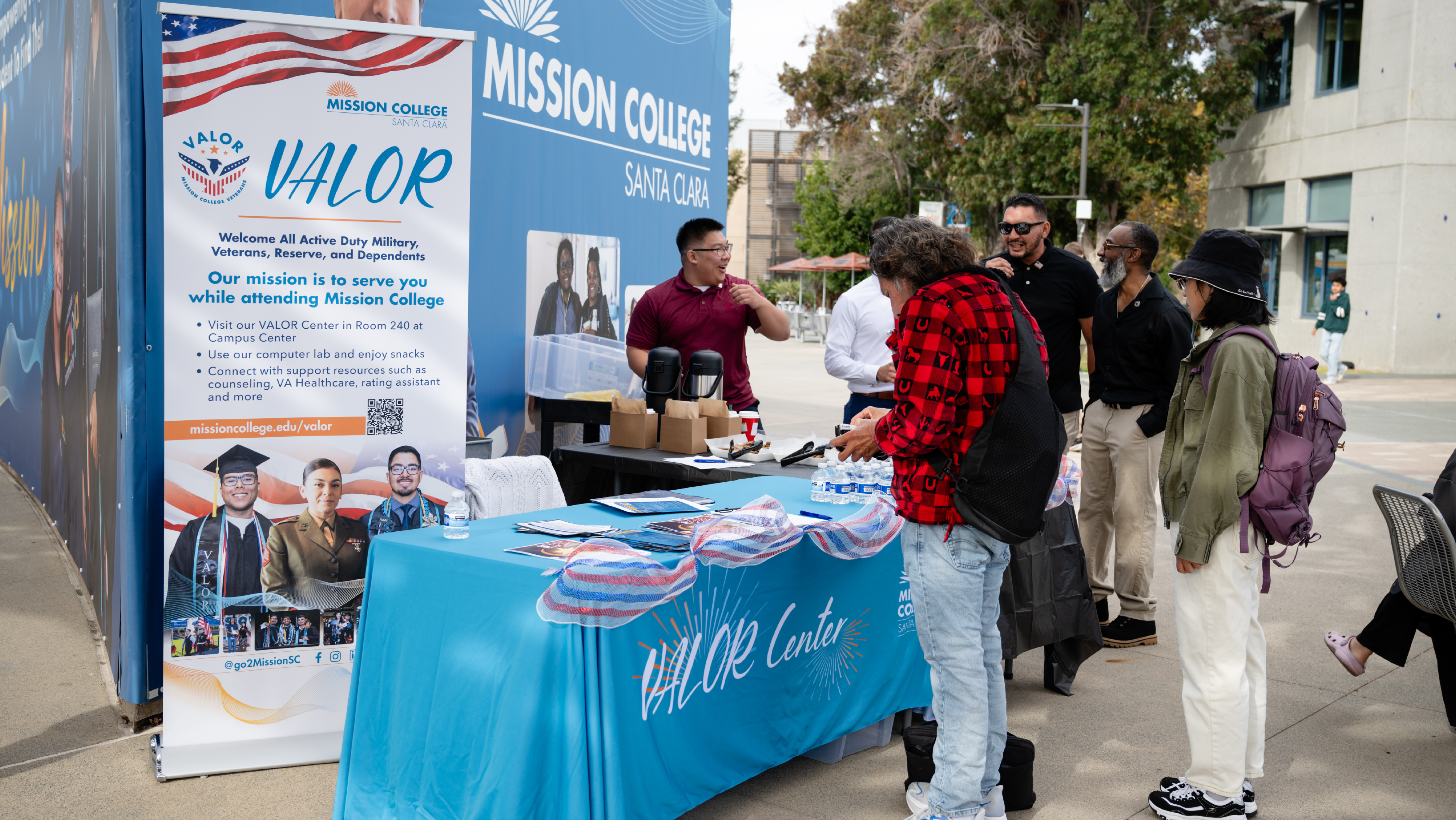 Students and visitors gathering around an outdoor Mission College VALOR Center information booth offering brochures and refreshments.
