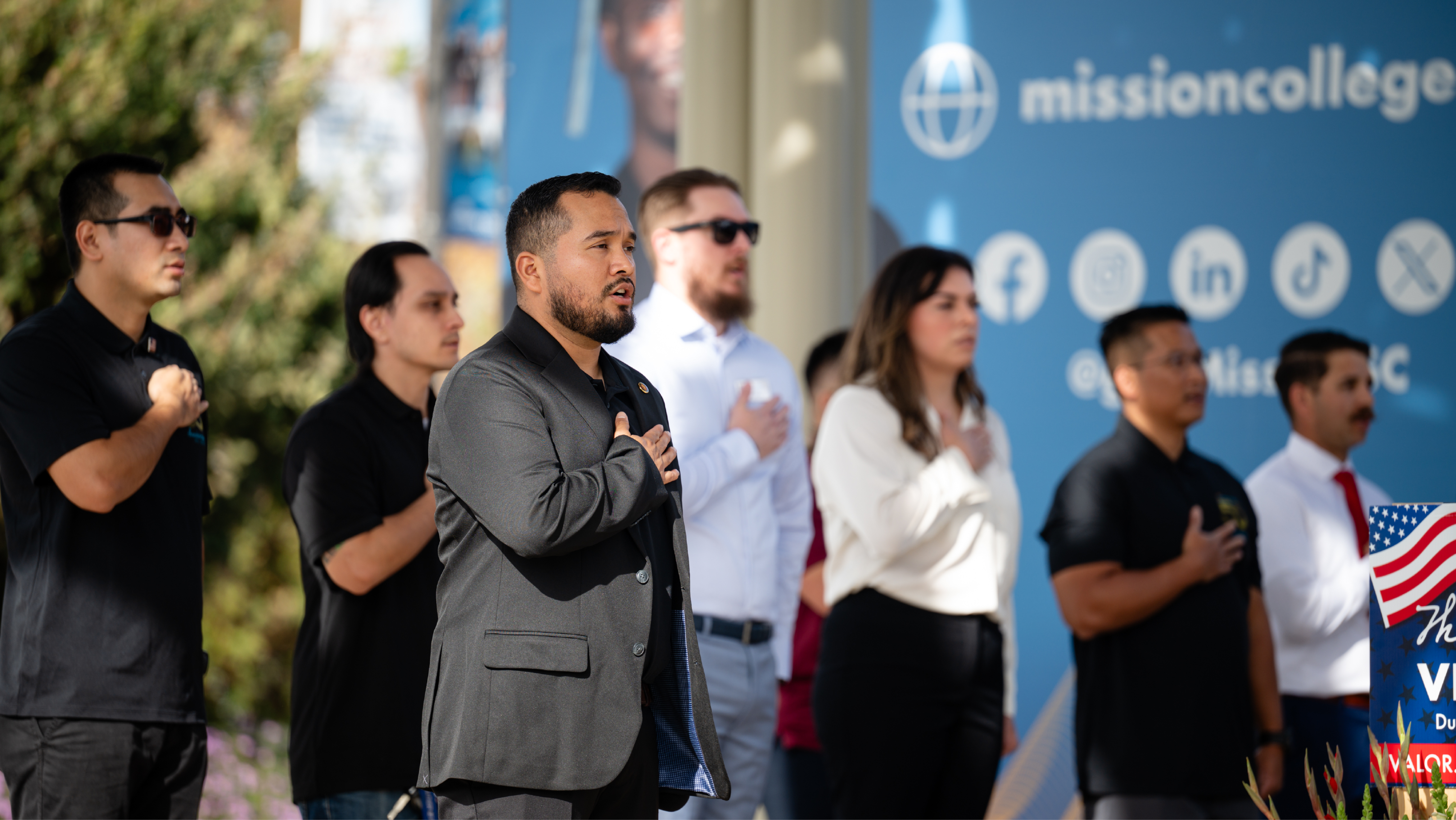 A row of attendees standing outdoors at a event with their right hands placed over their hearts during a ceremony.