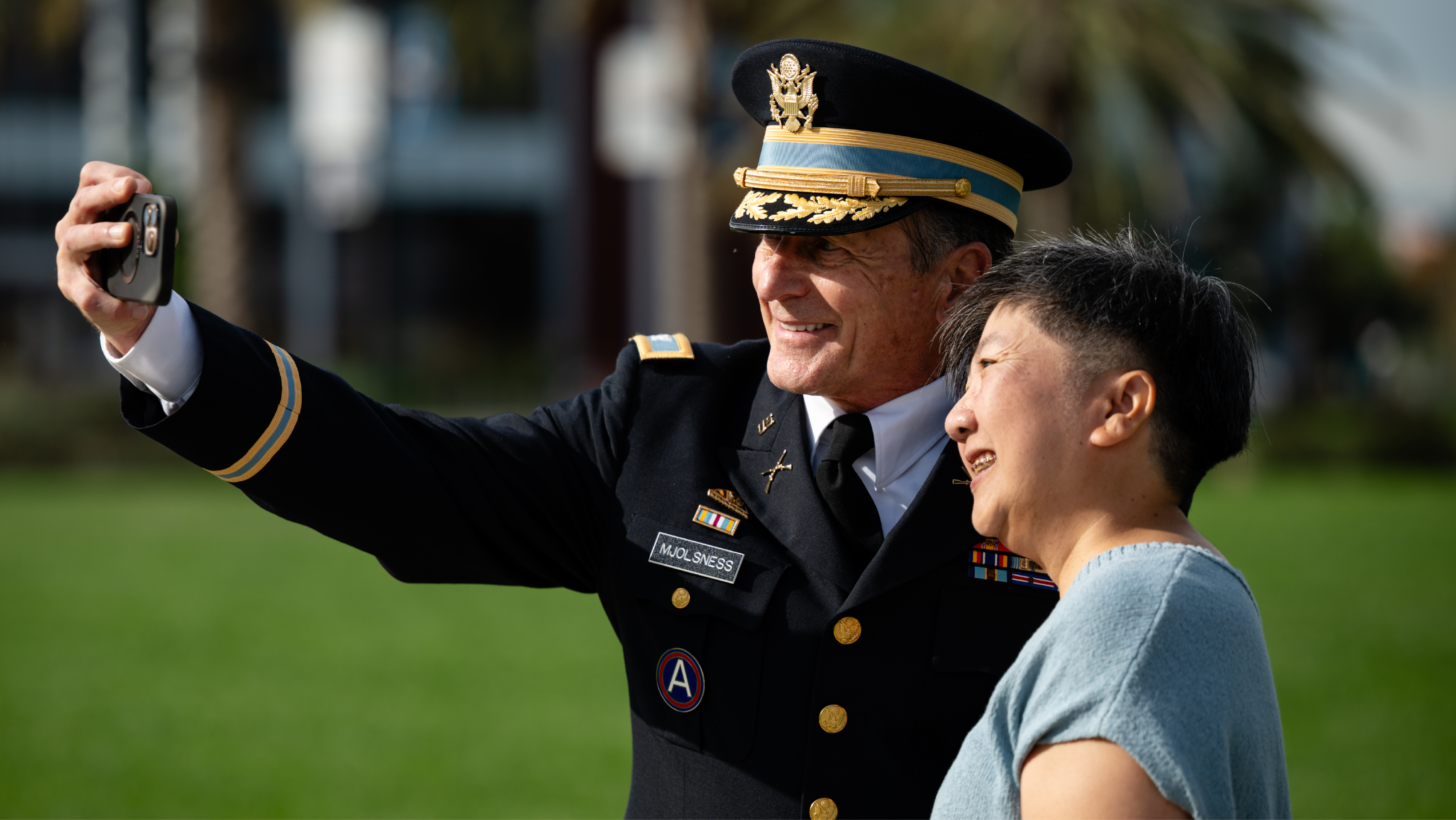 An Army officer in full dress uniform taking a selfie on a smartphone with a smiling woman on a sunny outdoor campus lawn.