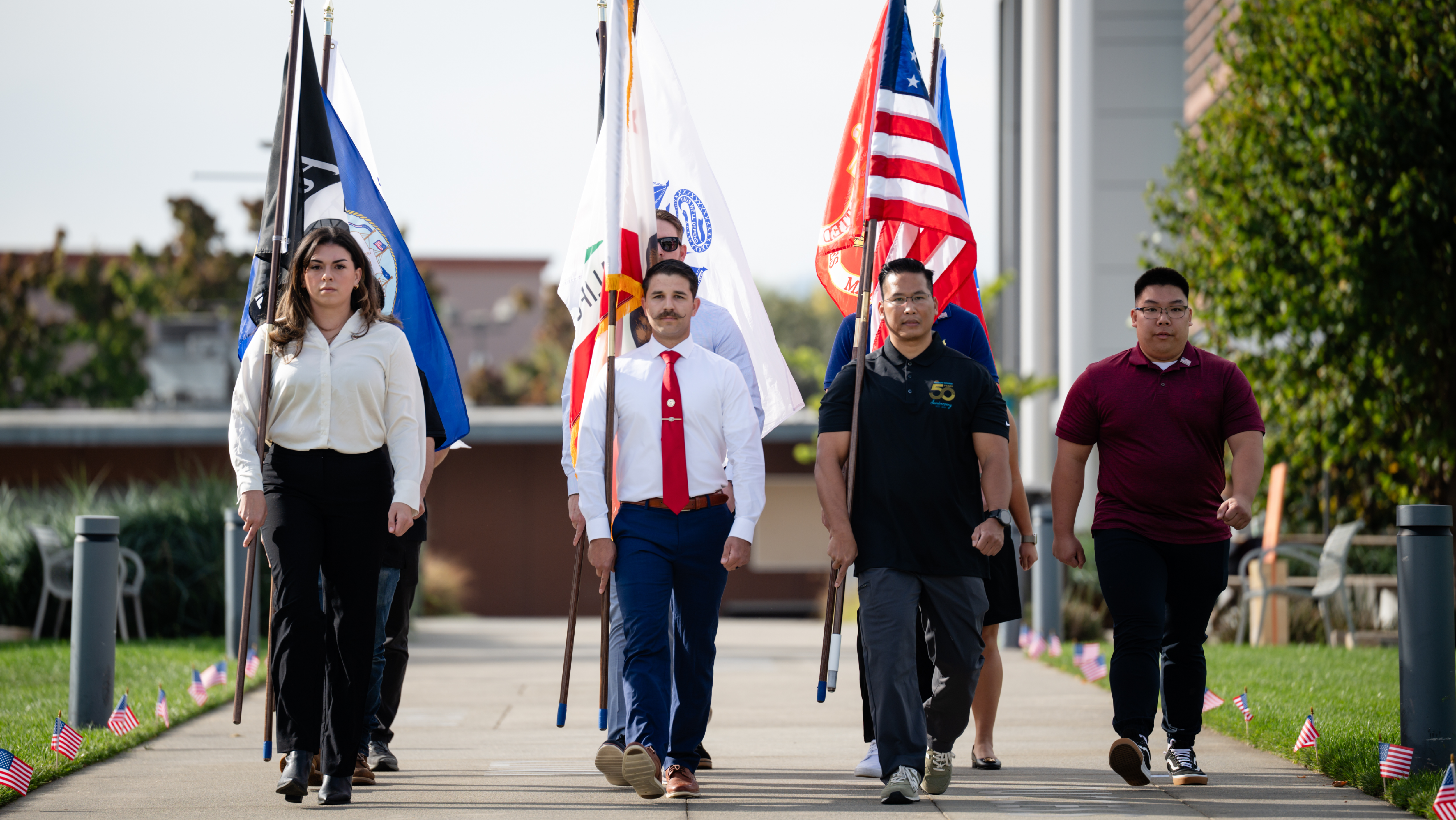A color guard unit carrying U.S. and military service flags down a outdoor concrete walkway lined with small American flags.